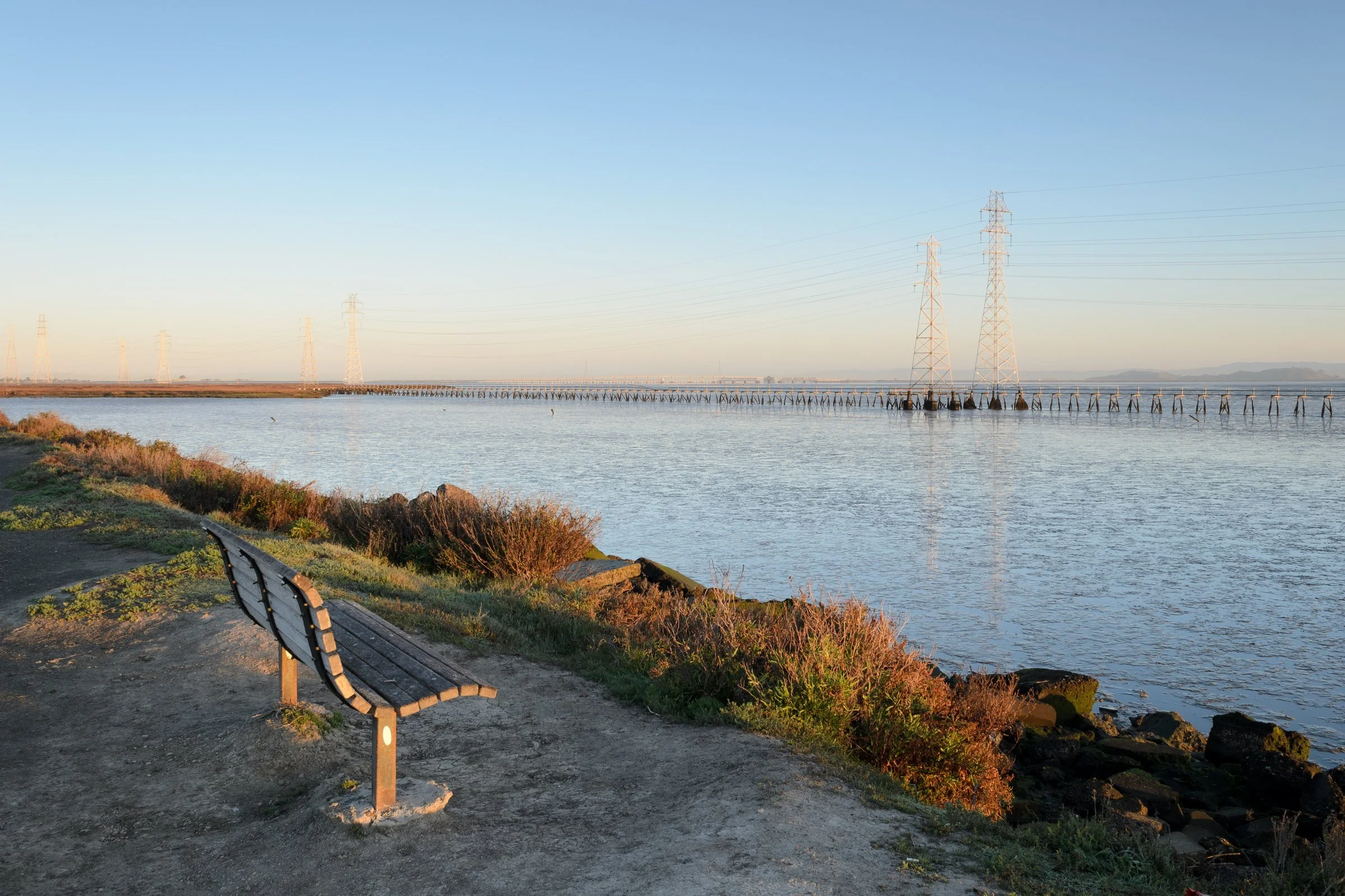 Dawn light over the Palo Alto Baylands, with a bench on a rocky shoreline, calm Bay water and a line of transmission towers on a long boardwalk.