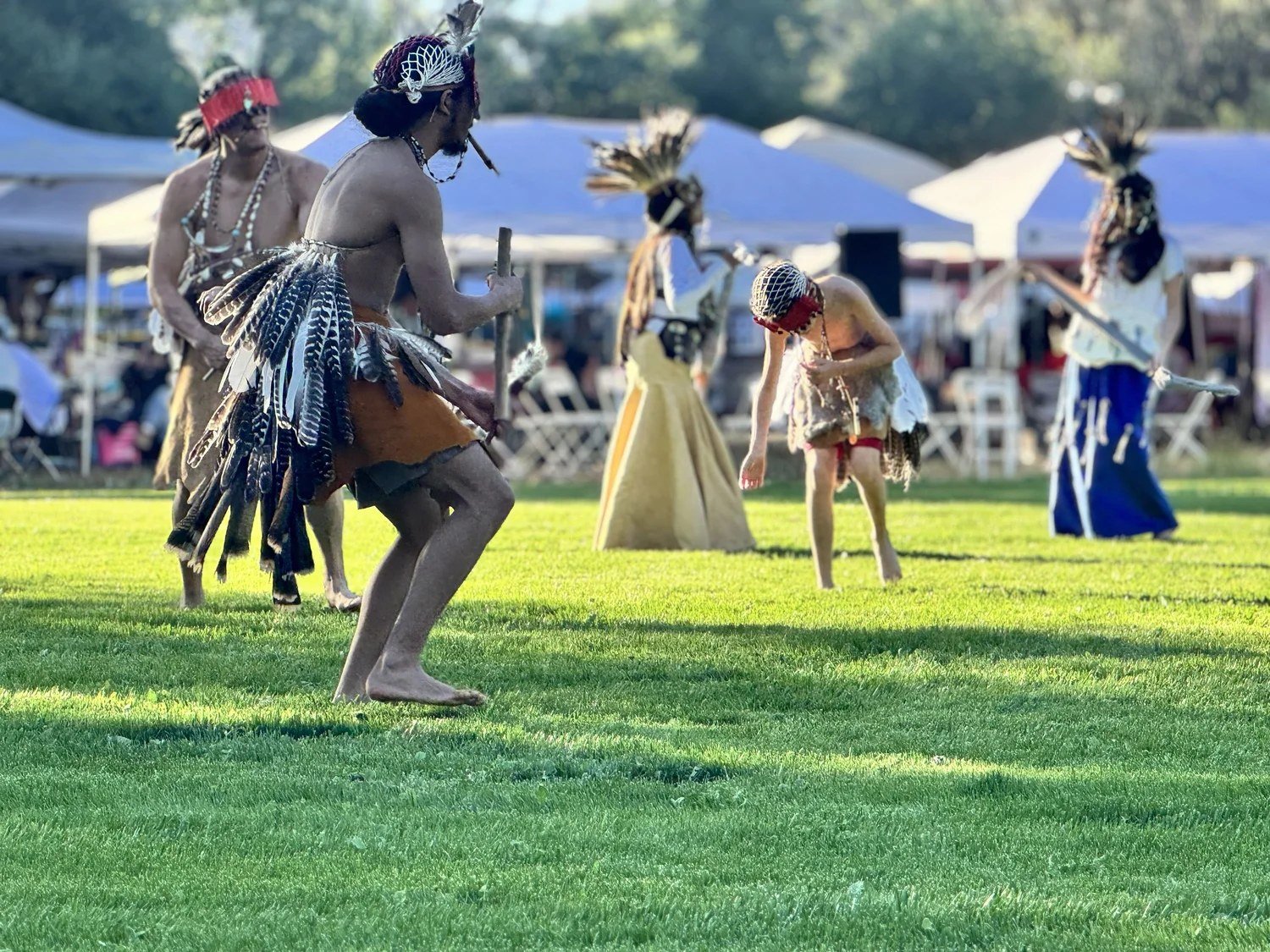 Dancers in feathered headdresses and regalia move across a green lawn at the Stanford Powwow, with tents and trees behind them.