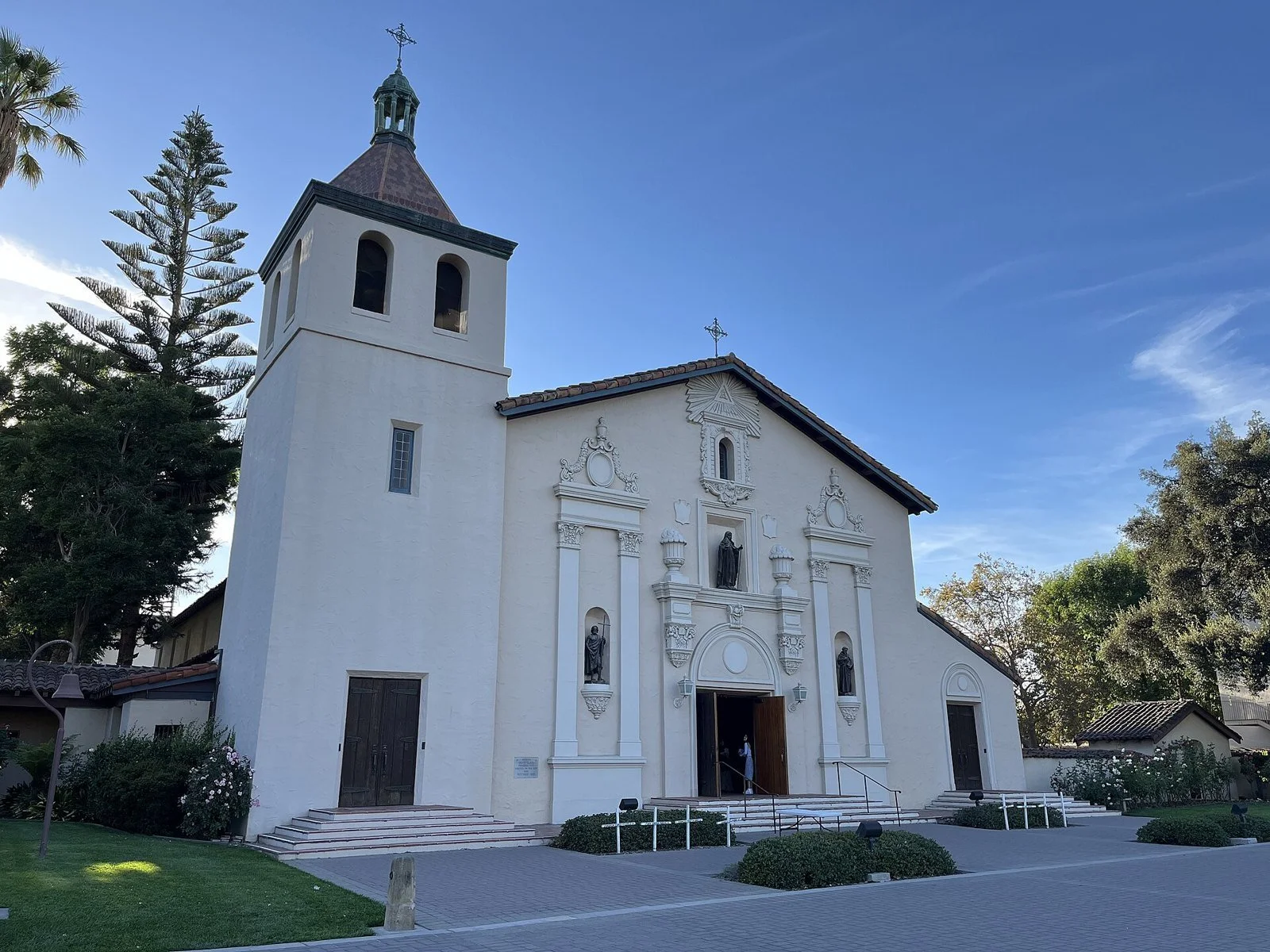 The painted facade and bell tower of Mission Santa Clara de Asís on the Santa Clara University campus, framed by trees.