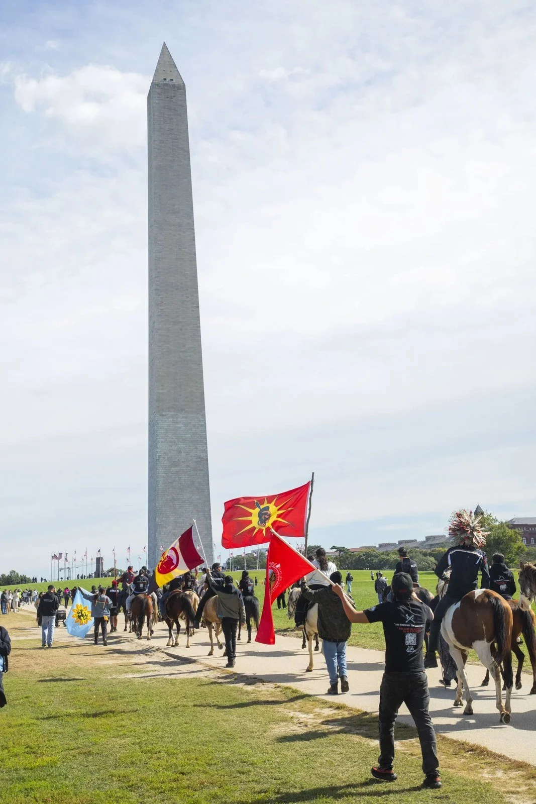 Riders on horseback and people on foot carrying red Muwekma Ohlone flags cross the grass of the National Mall with the Washington Monument behind them.
