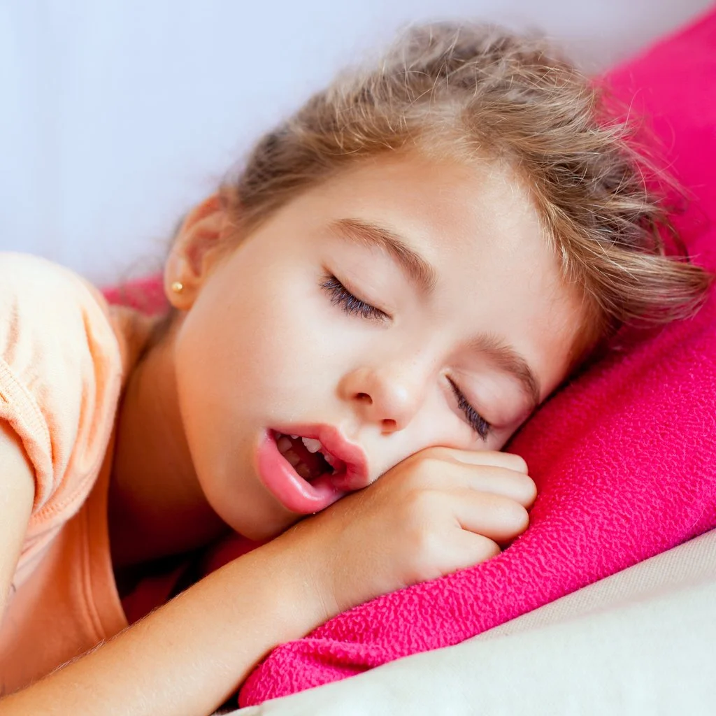 A young girl with light brown hair sleeping on a pink pillow with her hand near her face and her mouth slightly open.