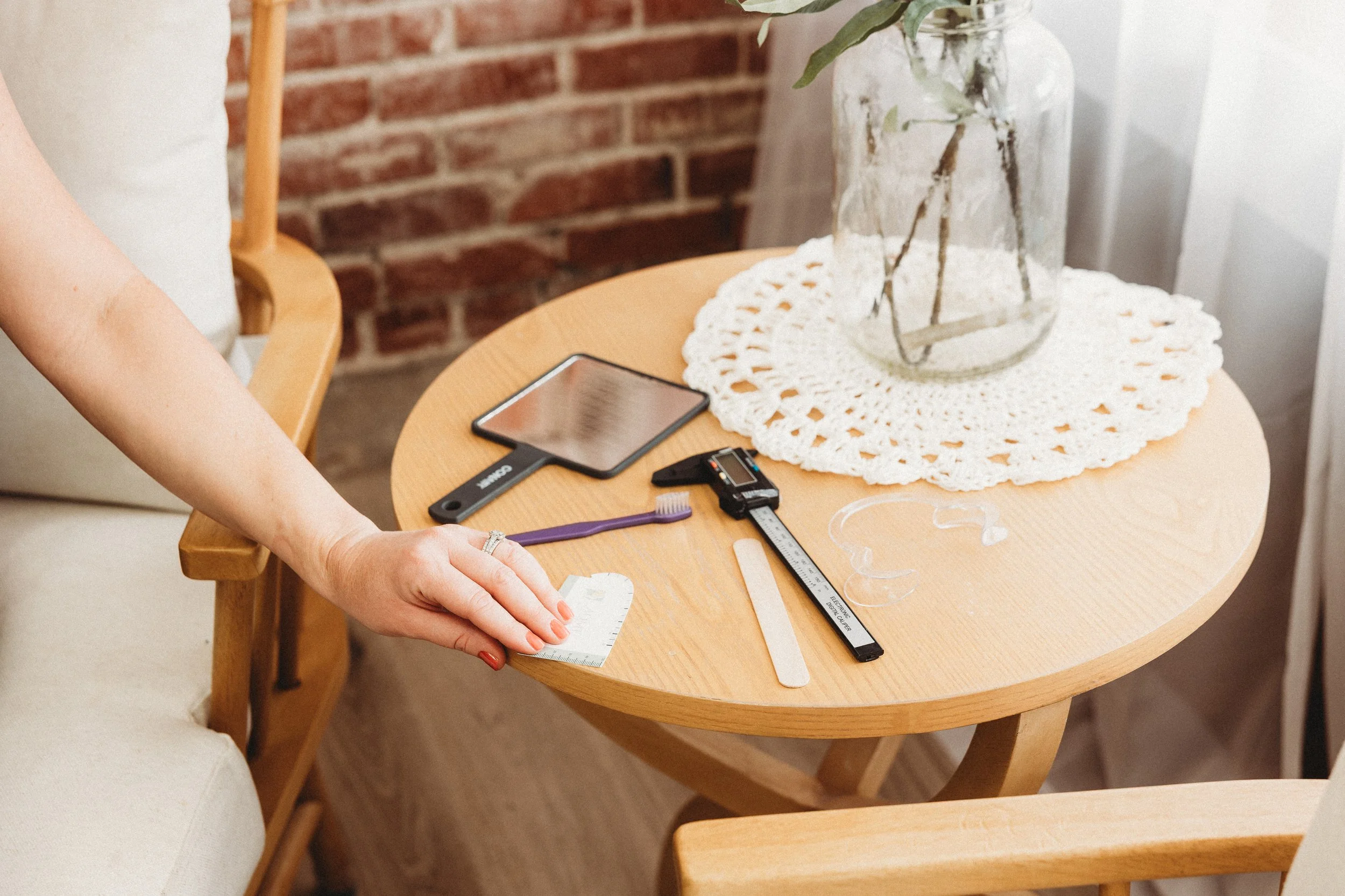 A person working with measuring tools on a small round wooden table, with a smartphone, a toothbrush, and a lace doily on the table, with a brick wall in the background.