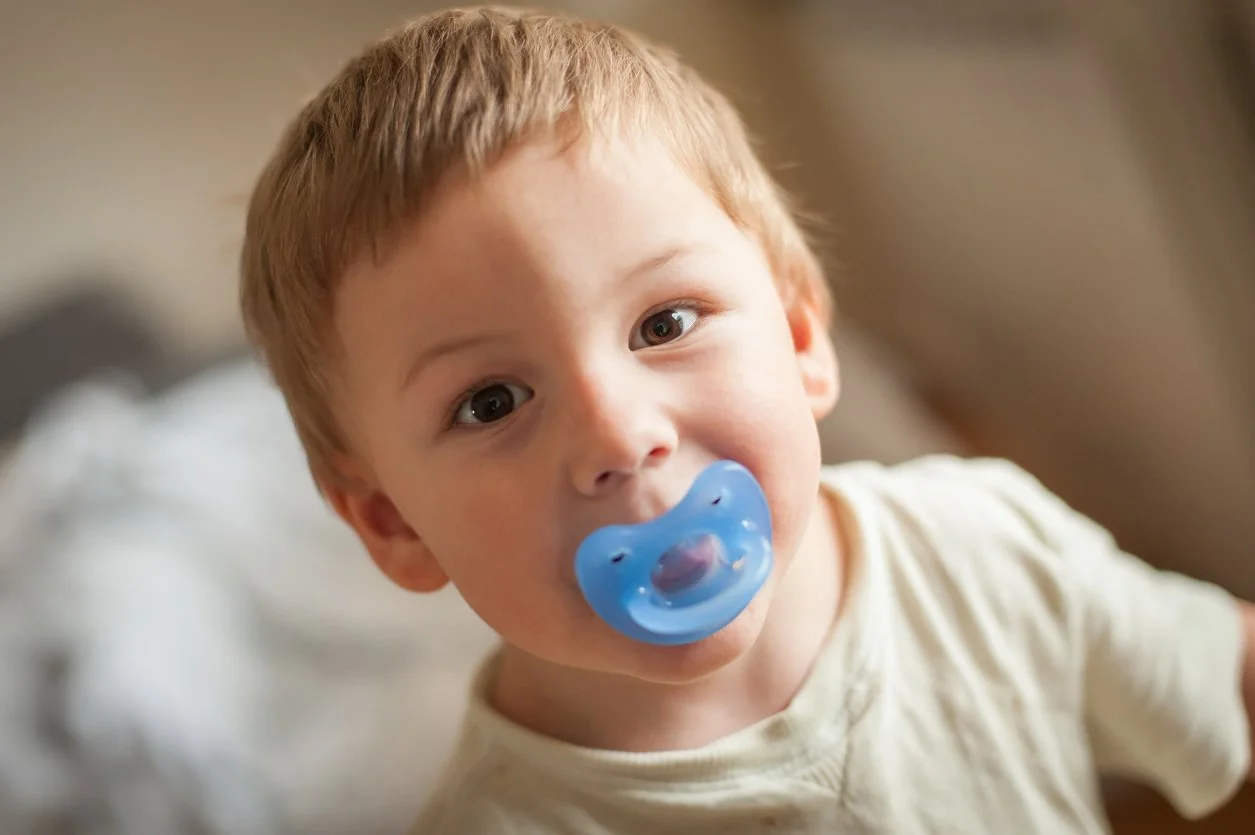 Close-up of a young boy with light brown hair and blue eyes, looking into the camera with a curious expression, holding a blue pacifier in his mouth.