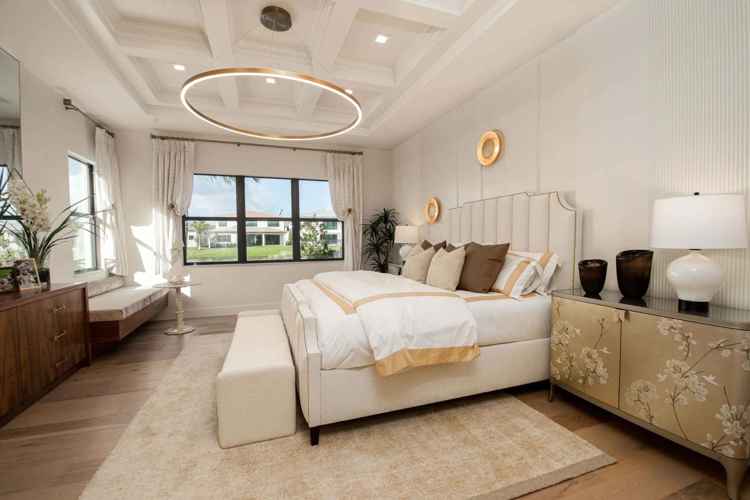 A modern bedroom with a large bed, beige and white bedding, a cream upholstered headboard, a floral-patterned sideboard, a white table lamp, and a circular ceiling light. There are windows with white curtains, a wooden dresser with photo frames, and