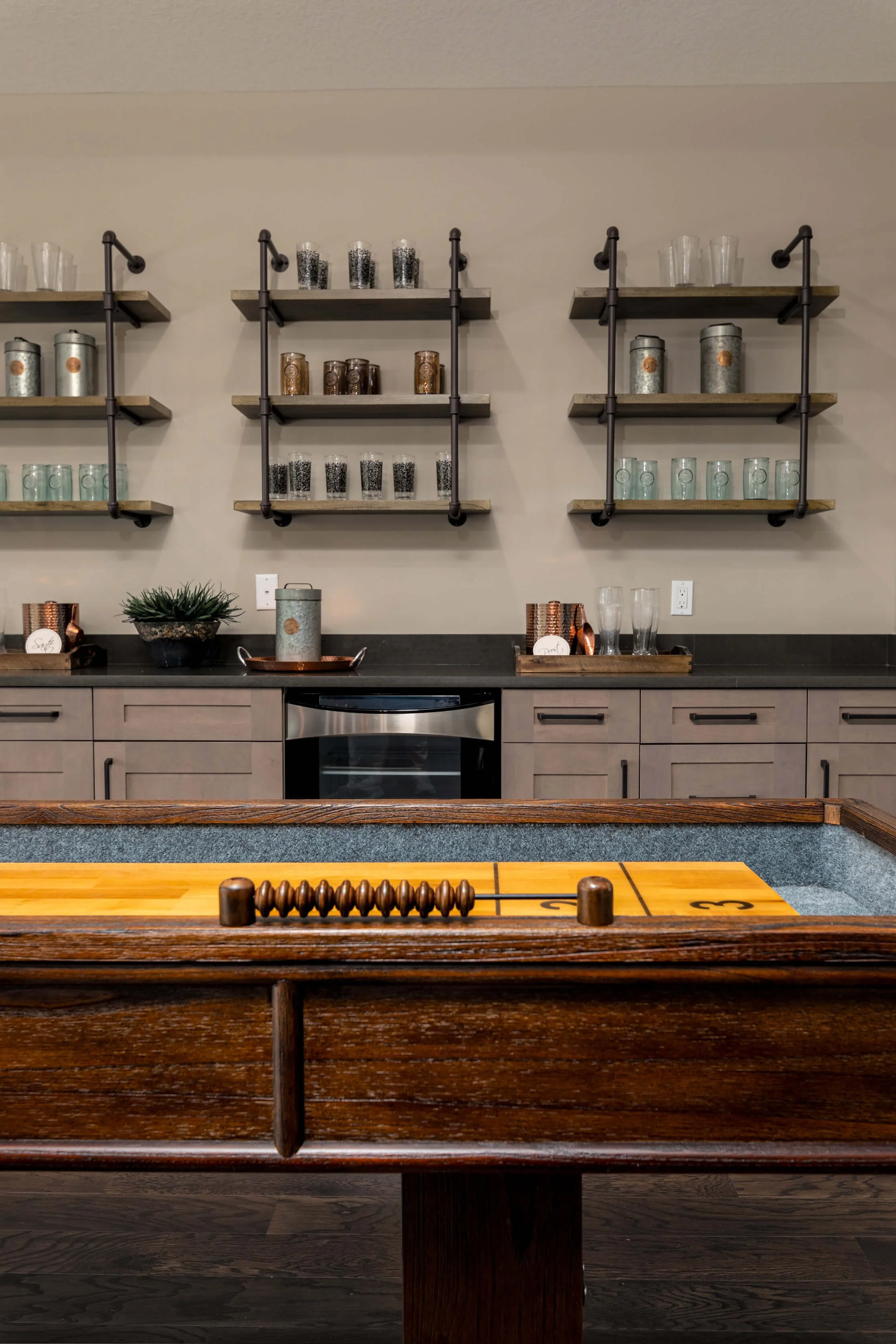 Interior of a game room with a shuffleboard table in the foreground and a kitchen-style backdrop with open shelves, glassware, and decorative items.