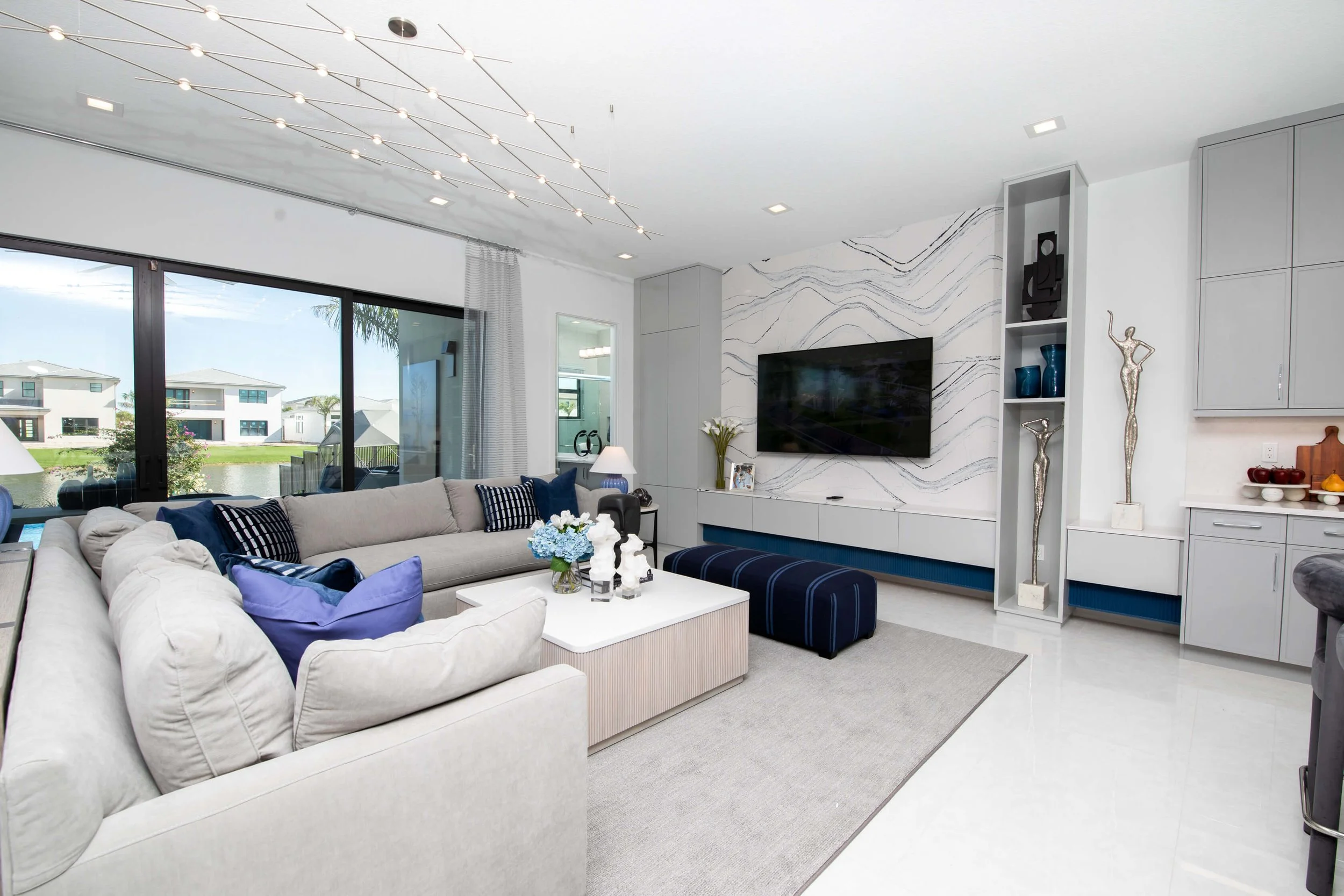 Modern living room with large gray sectional sofa, navy and striped cushions, white coffee table with decorative items, gray area rug, wall-mounted flat-screen TV, white cabinetry, decorative sculptures, and large sliding glass doors showing a view o