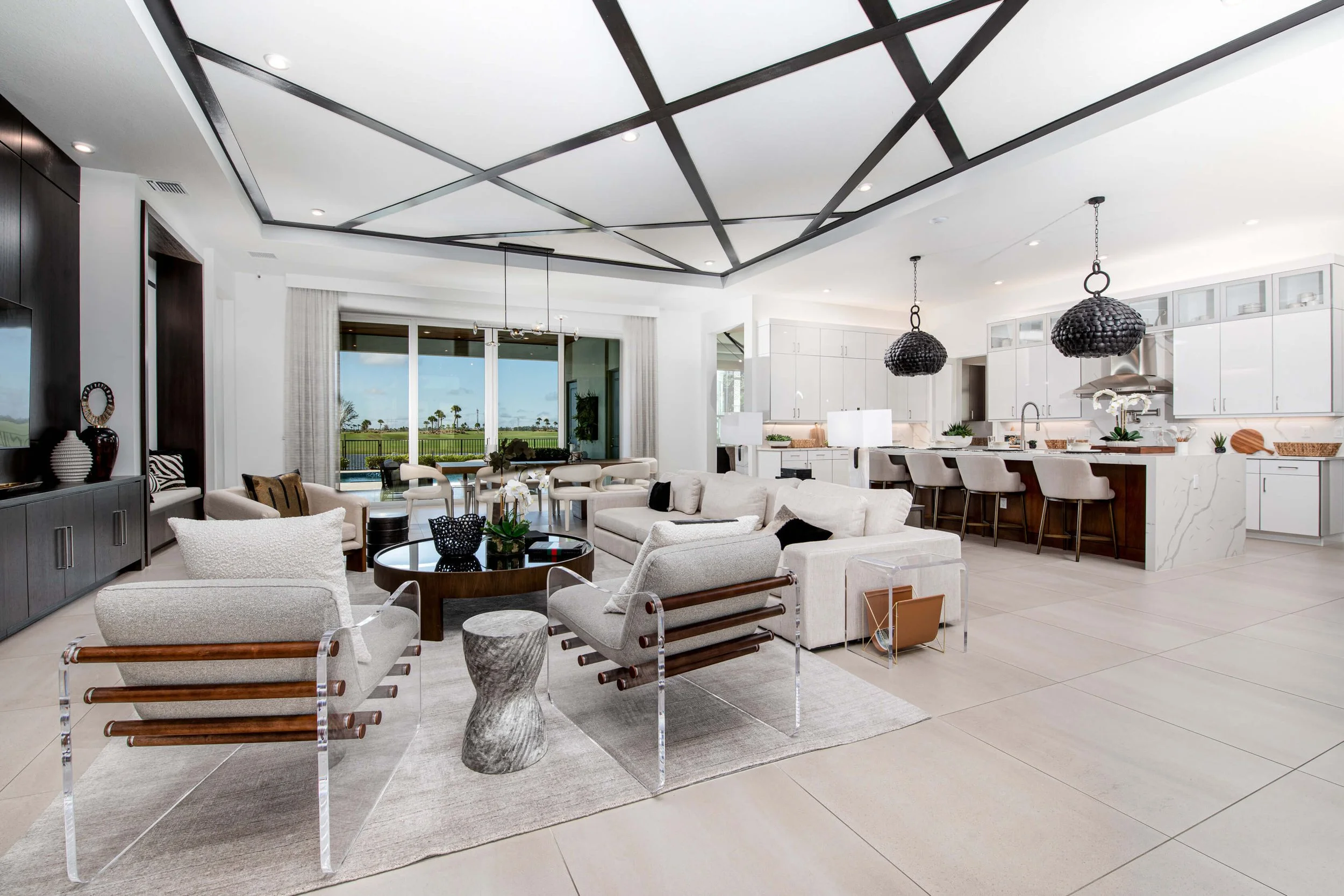 Open-concept living room and kitchen with modern white cabinets, a marble kitchen island, beige chairs, and black pendant lights, with large sliding doors leading to an outdoor space.