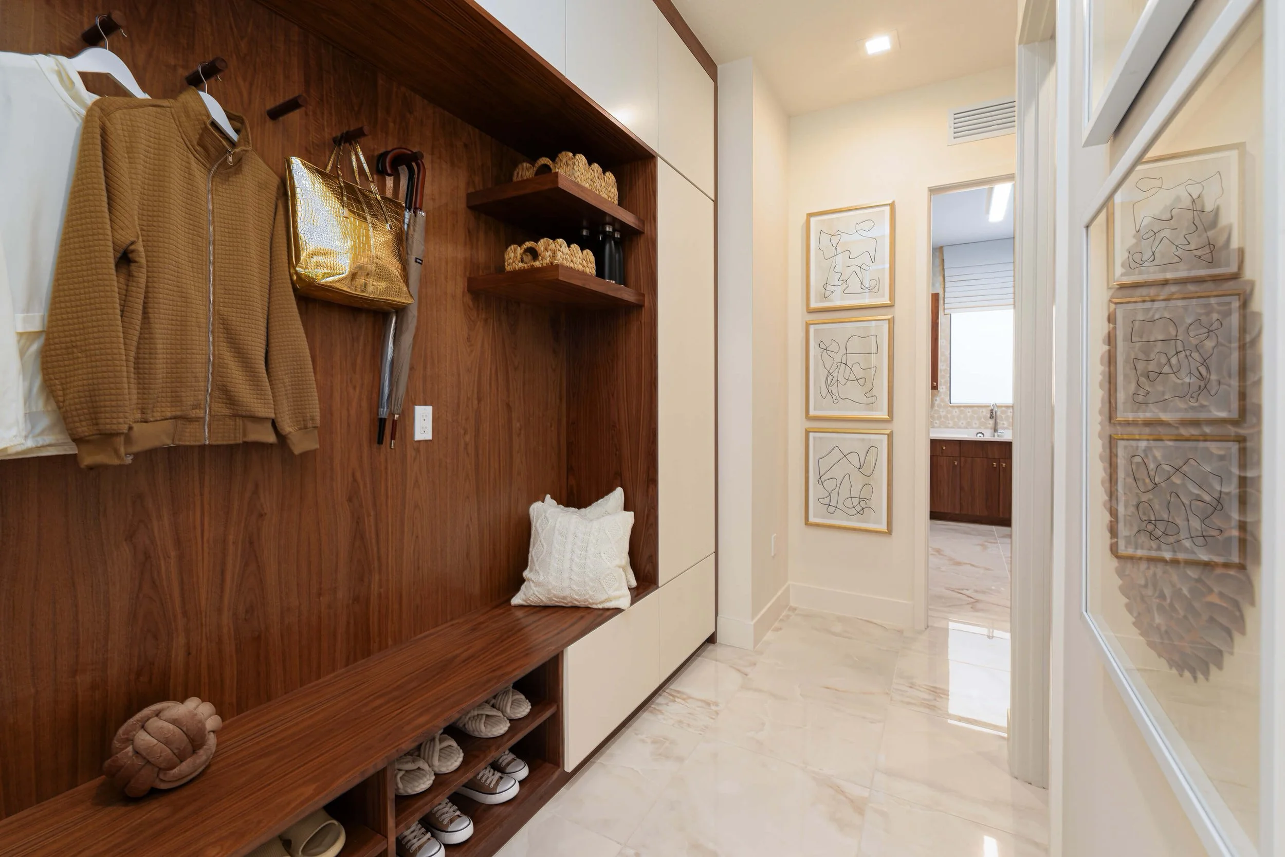 Entryway with wood panel wall, hanging clothes, shelves with decor, framed abstract art, marble floor, and part of a kitchen visible in the background.