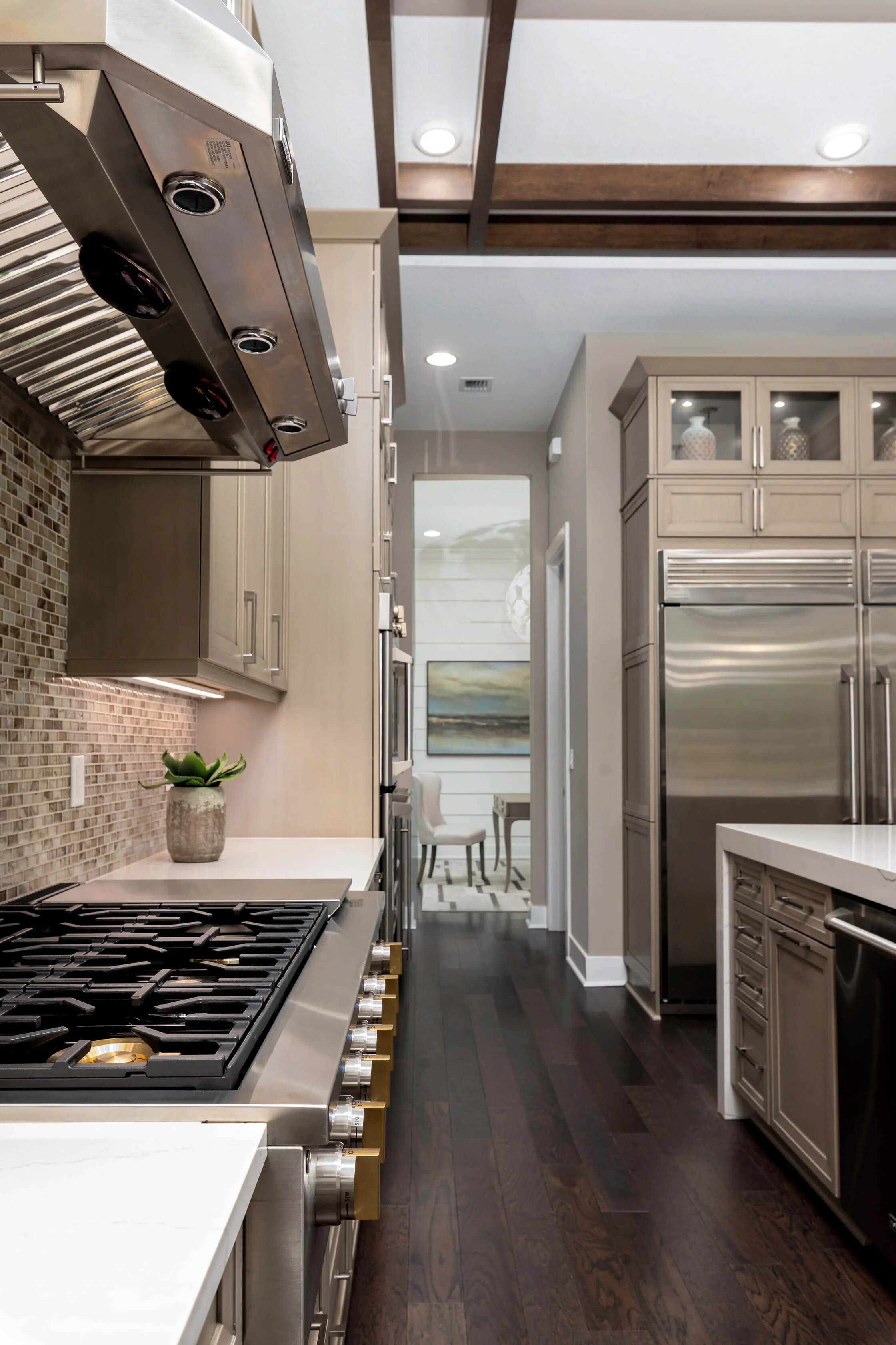 Modern kitchen with stainless steel appliances, beige cabinetry, dark hardwood floors, and a view into a dining area with chairs and artwork.