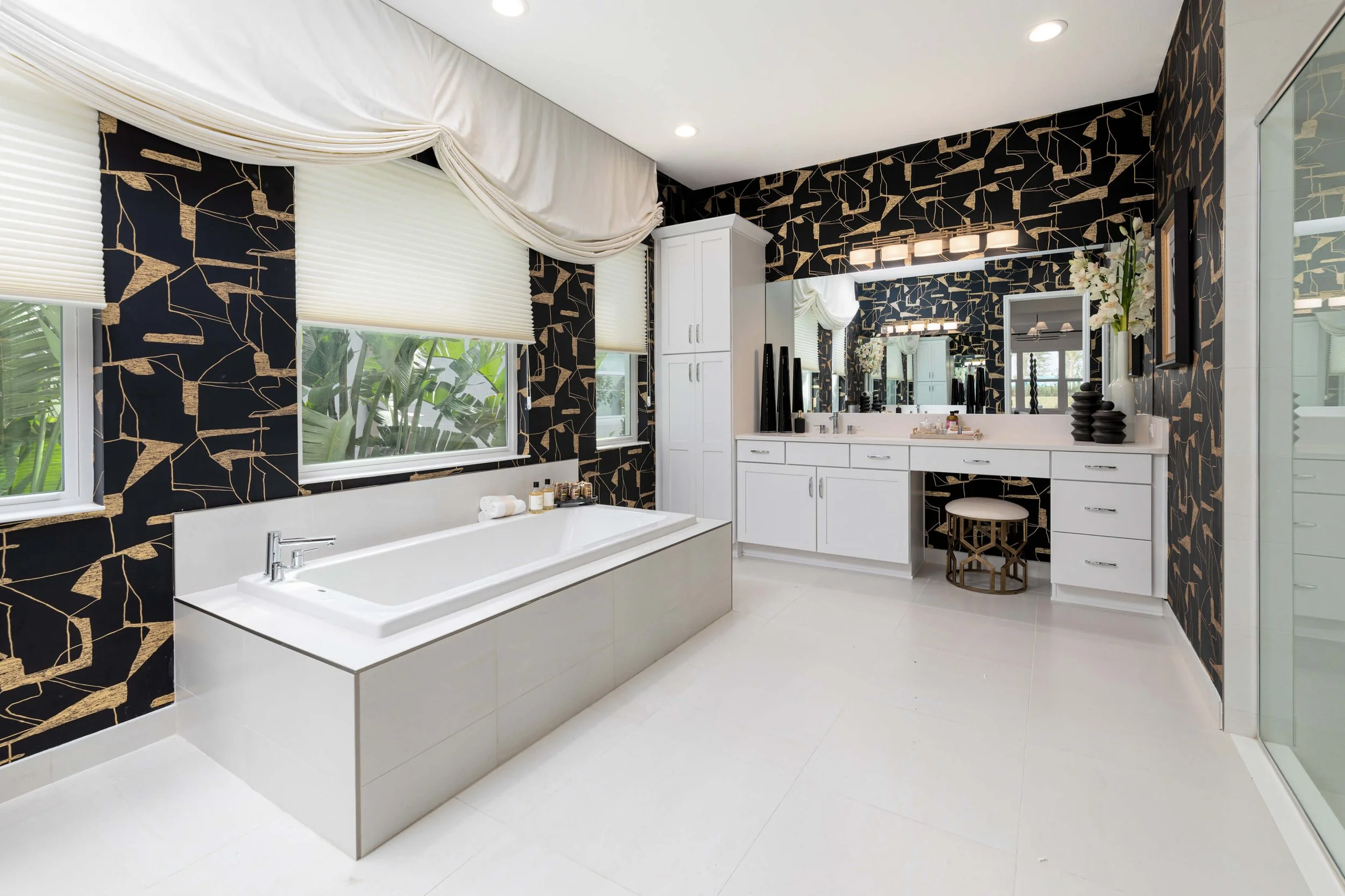 Modern bathroom with black and gold patterned wallpaper, a large bathtub, white cabinets, a vanity with mirror, and sliding glass door.