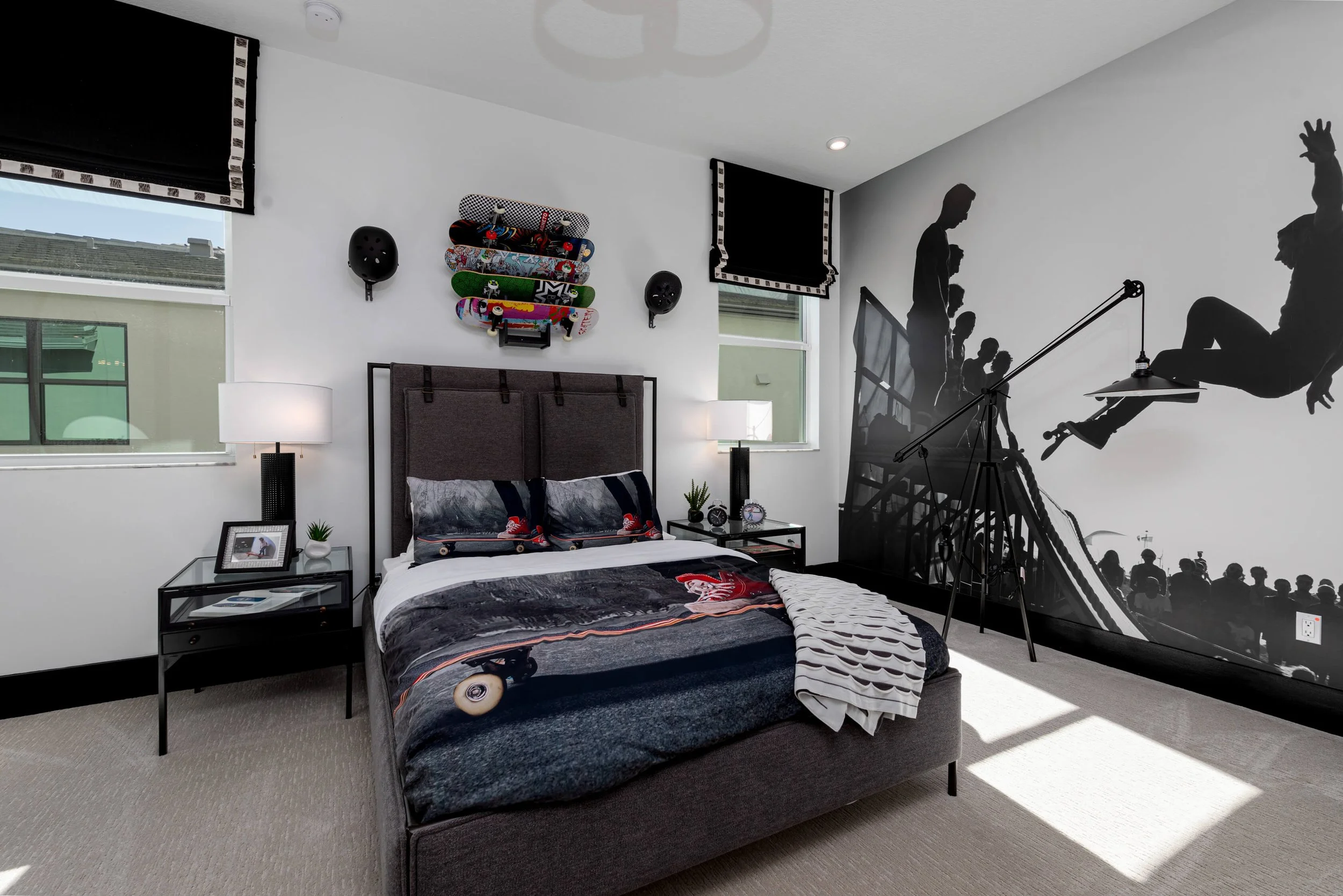 Bedroom with skateboard-themed bedding, skateboard wall art, black wall mural of skateboarders, and a bicycle silhouette mural, with two windows, nightstands with lamps, and beige carpet.