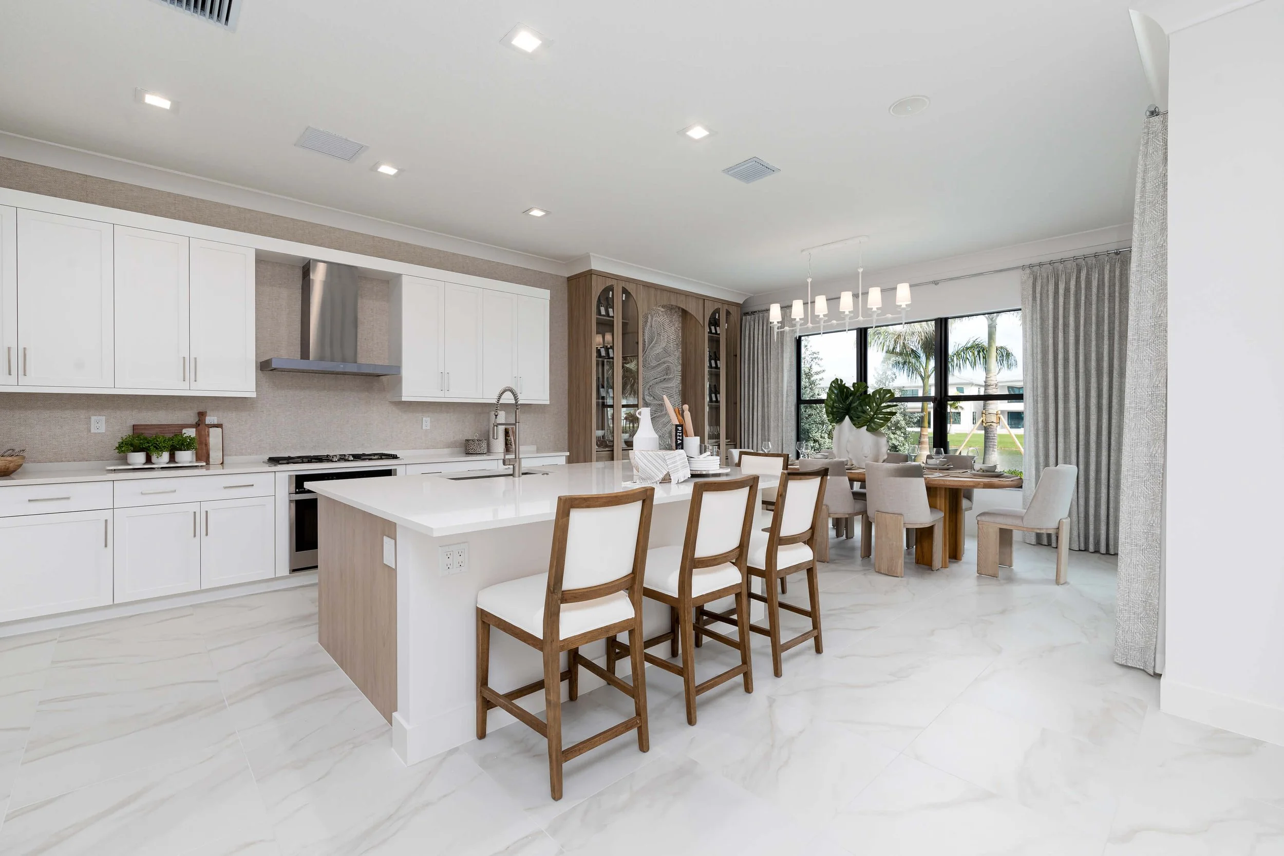 Modern kitchen and dining area with white cabinets, kitchen island, and wooden dining table with chairs, large windows showing outdoor palm trees, light-colored tiled flooring, and neutral decor.