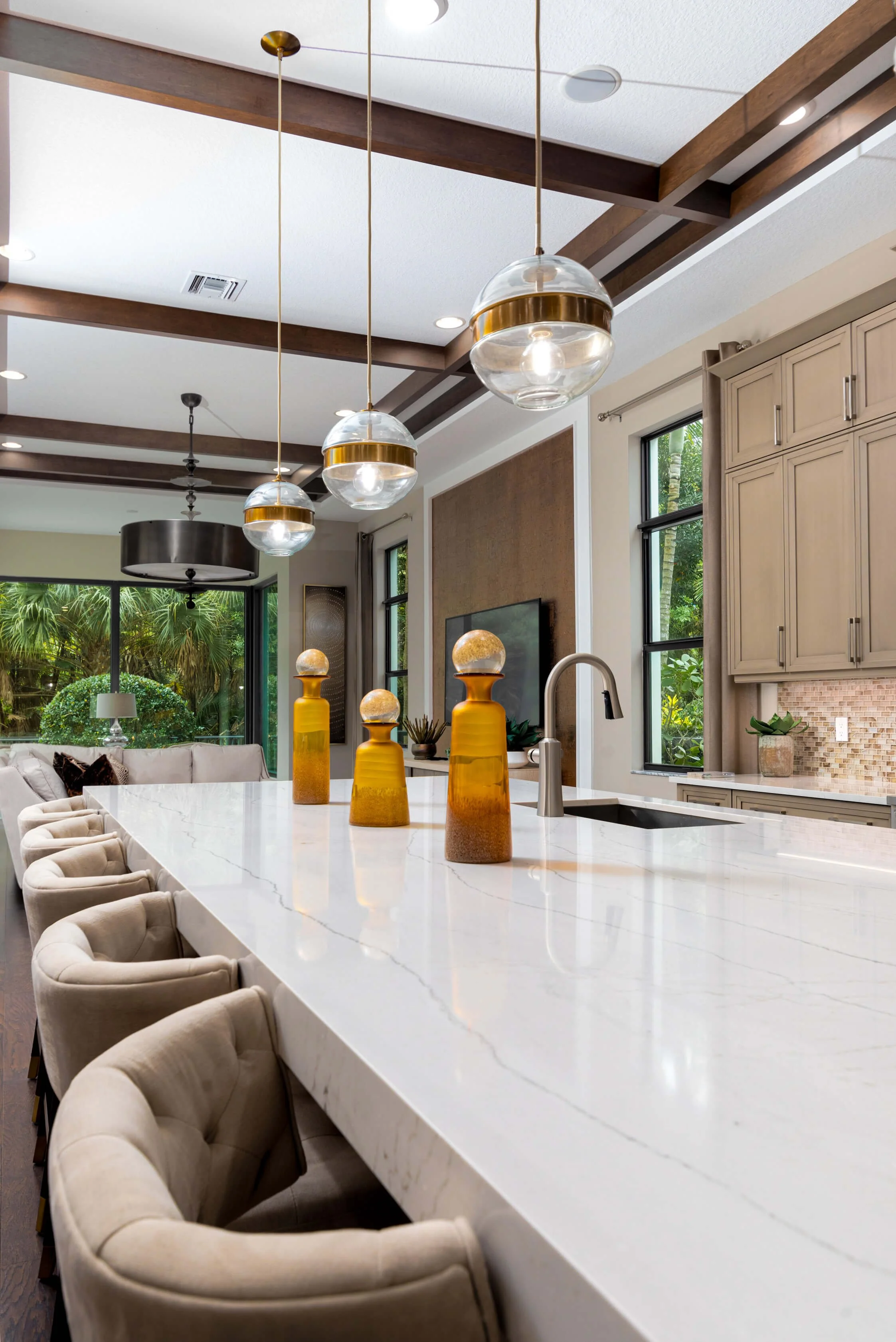 Modern kitchen with white marble island, pendant lights, beige chairs, large windows, and a view of outdoor greenery.