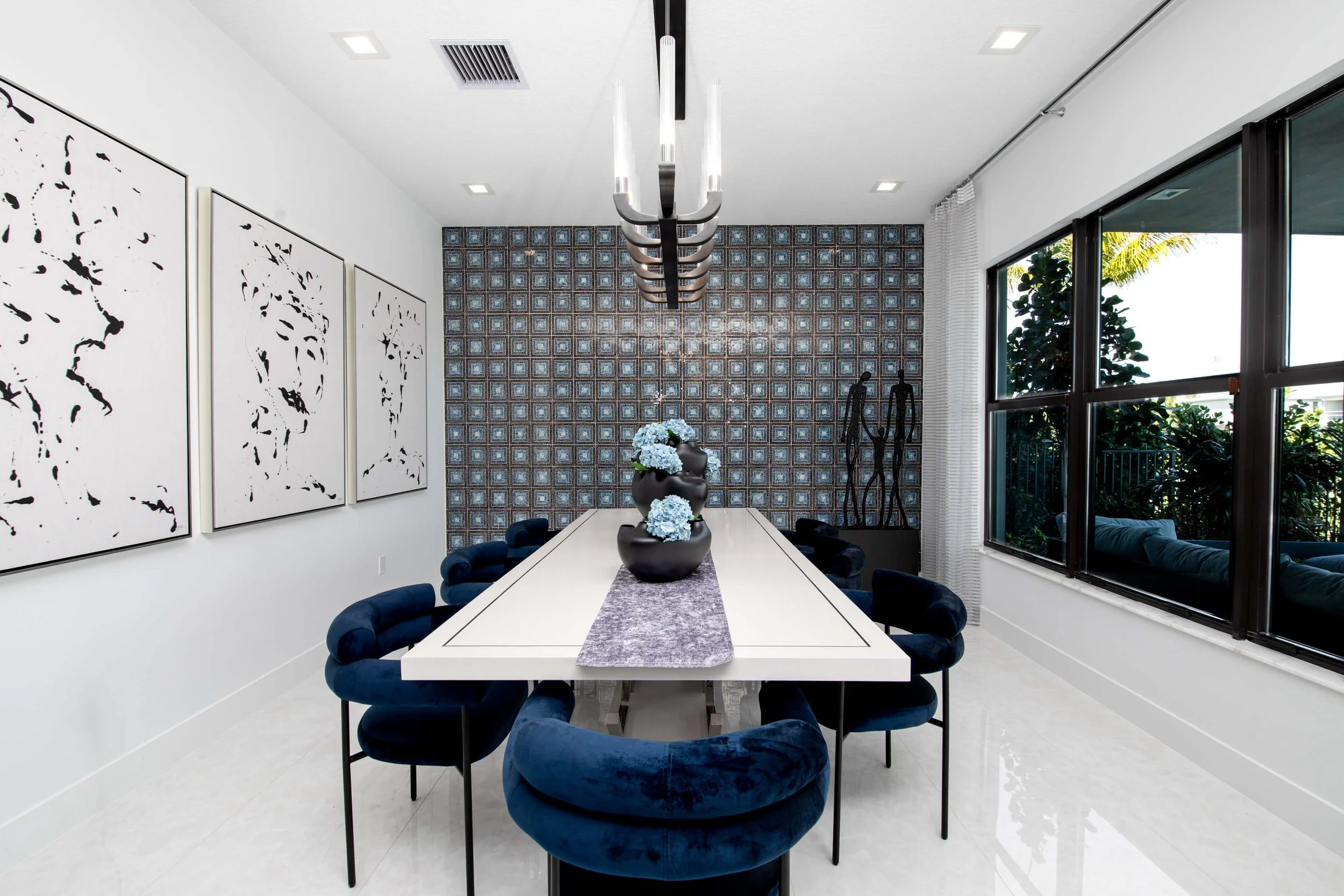 Modern dining room with white table, blue velvet chairs, black vases with blue flowers, wall art, large windows showing outdoor greenery, and a decorated accent wall.