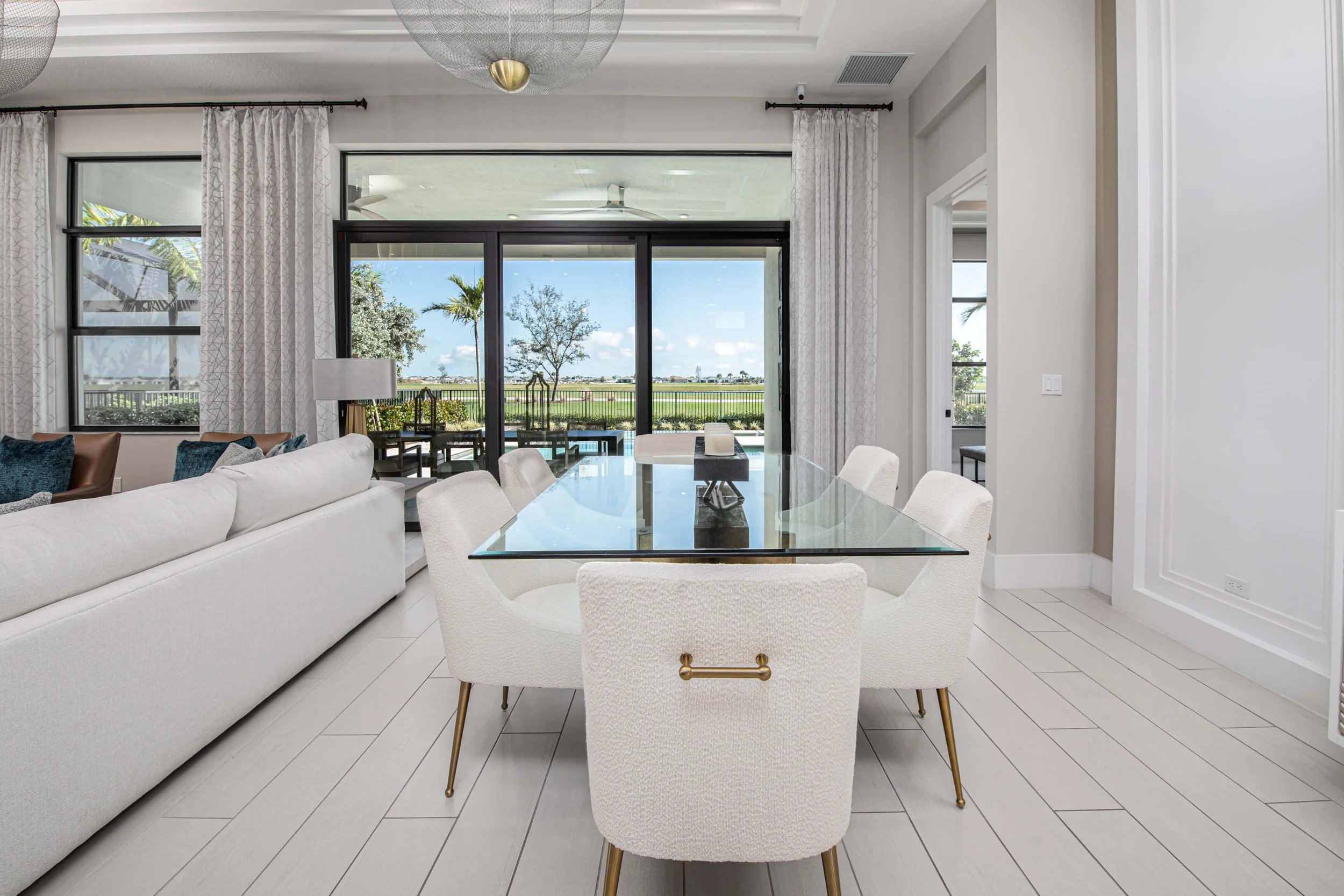 Modern dining area with a glass table, white upholstered chairs, and a view of a backyard with trees and a pool through sliding glass doors.