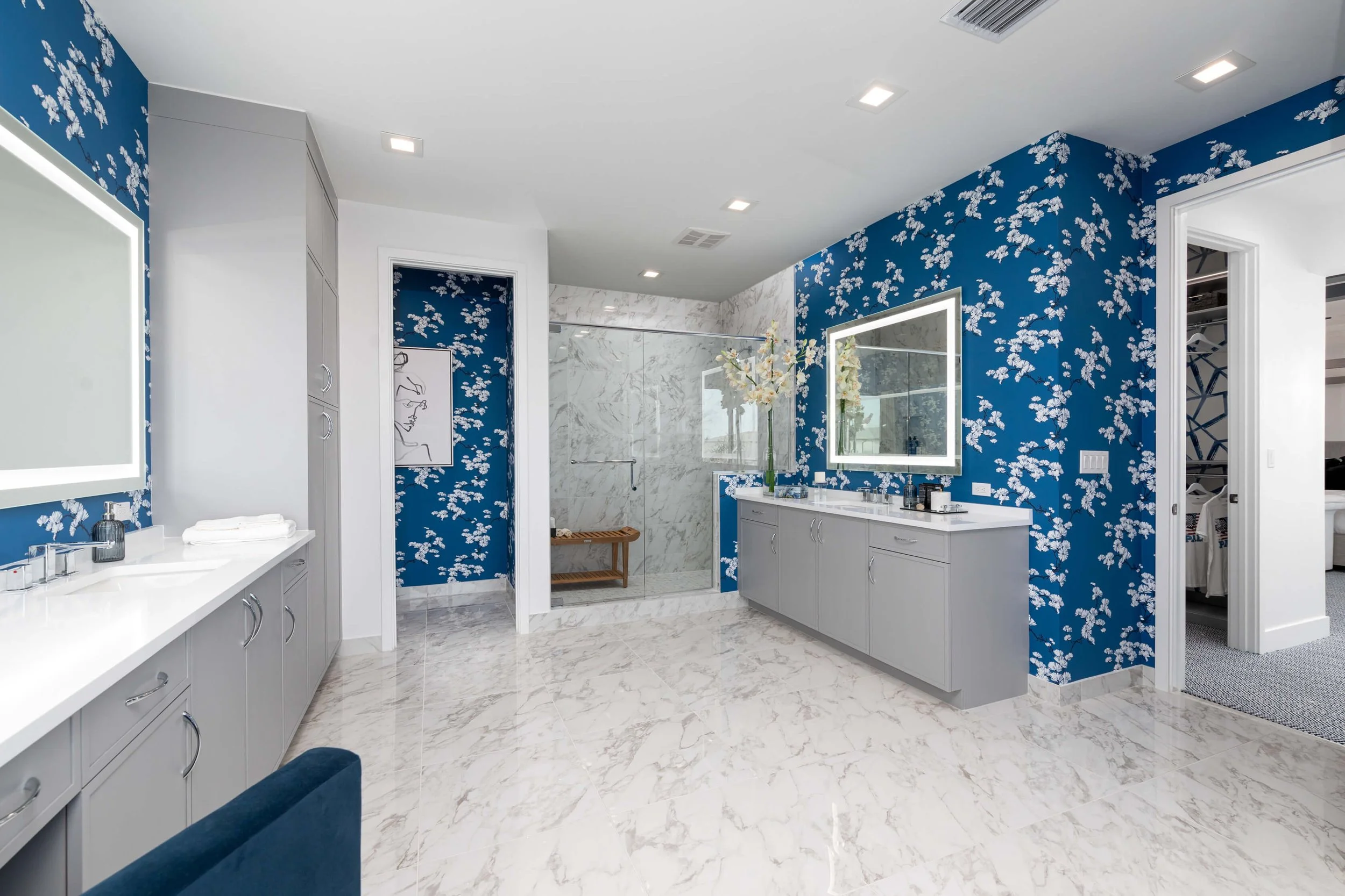 Modern bathroom with blue floral wallpaper, white marble floors, a double vanity with gray cabinets, and a glass-enclosed shower with white marble walls.