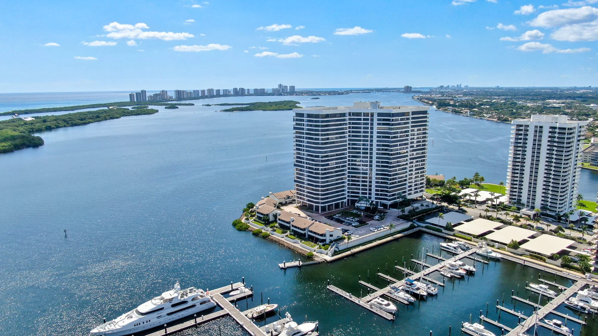 High angle aerial view of a marina with yachts, a large waterfront apartment building, and a river with lush landscape and cityscape in the background.