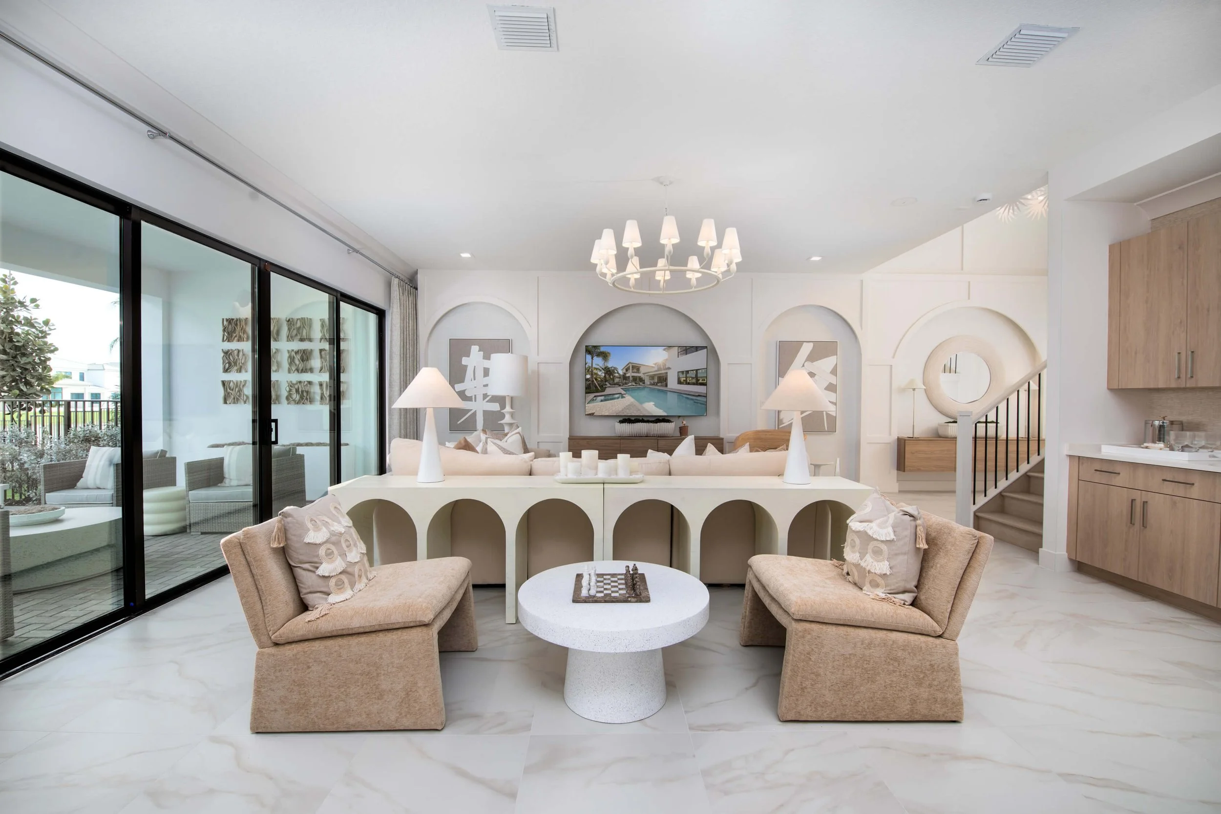Modern living room with beige chairs, a white curved sofa, a round coffee table with a chess set, sliding glass doors leading to an outdoor patio, and a wall-mounted television.
