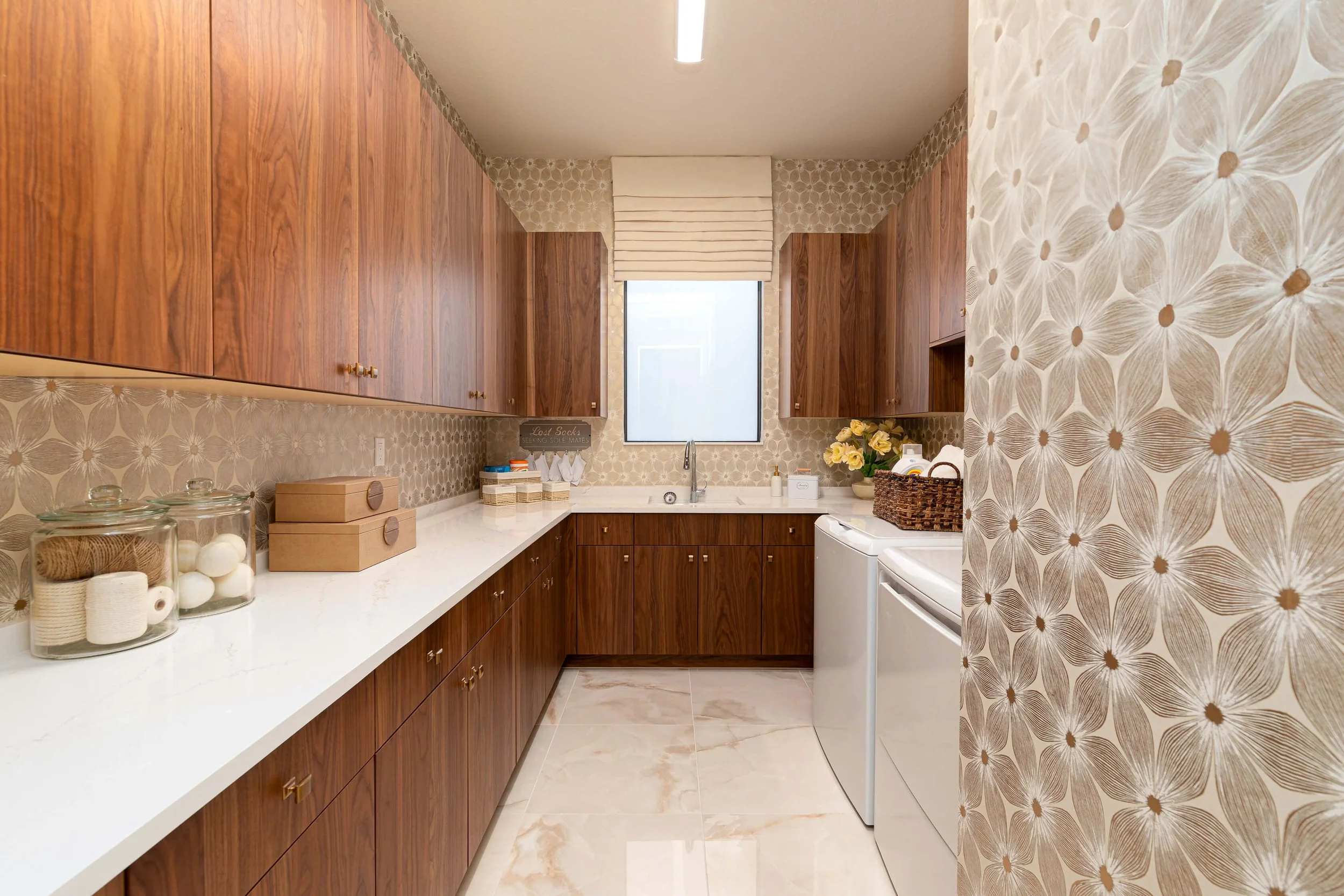 A laundry room with wood cabinets, a white countertop, a stainless steel sink under a window, a washer and dryer, and decorative wall and backsplash patterns.