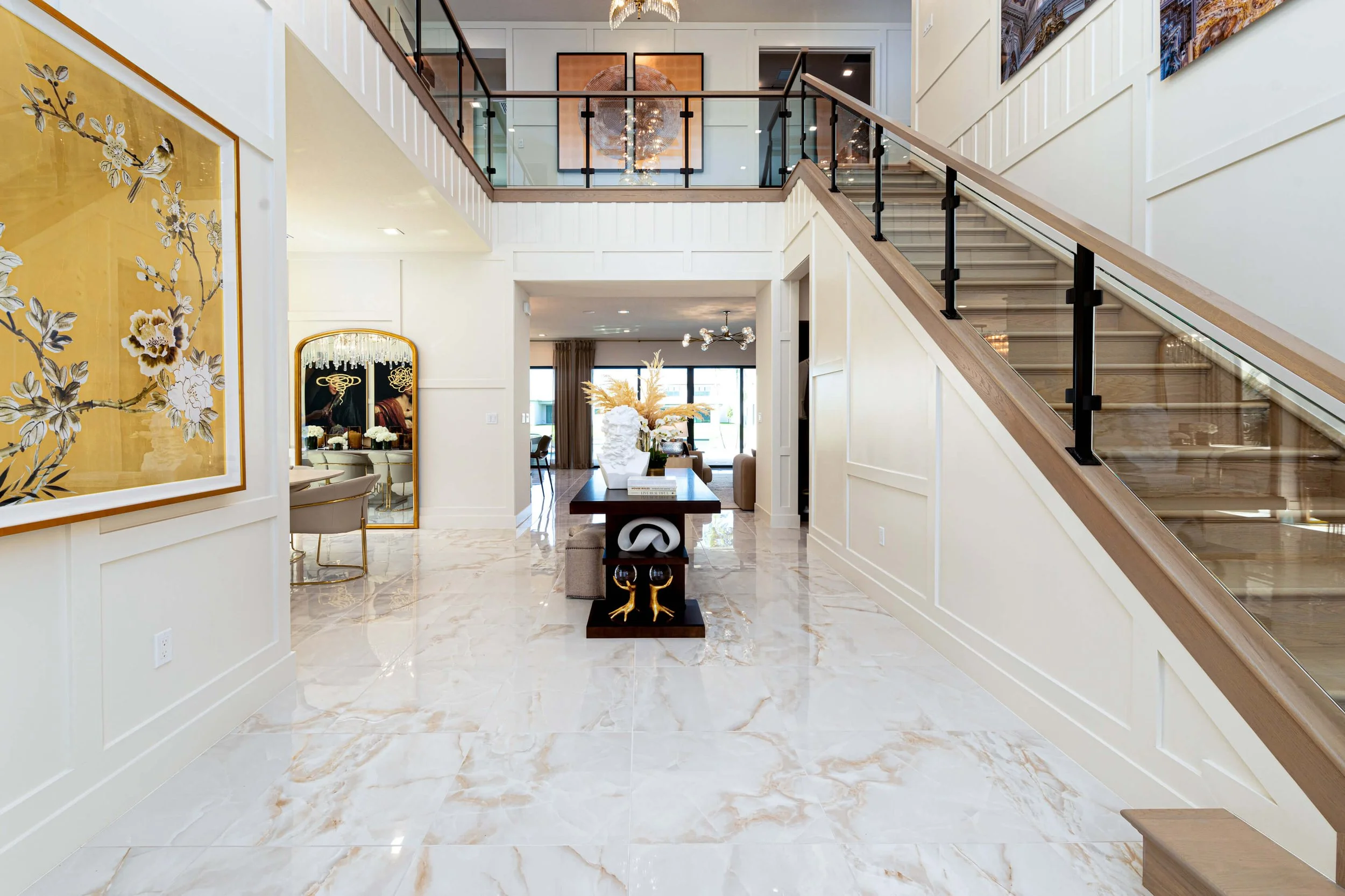 Interior of a luxury home, bright living space with marble floors, staircase with glass and wood railing, artwork on the walls, a table with decorative objects, and view into another room with dining area and outdoor scenery.
