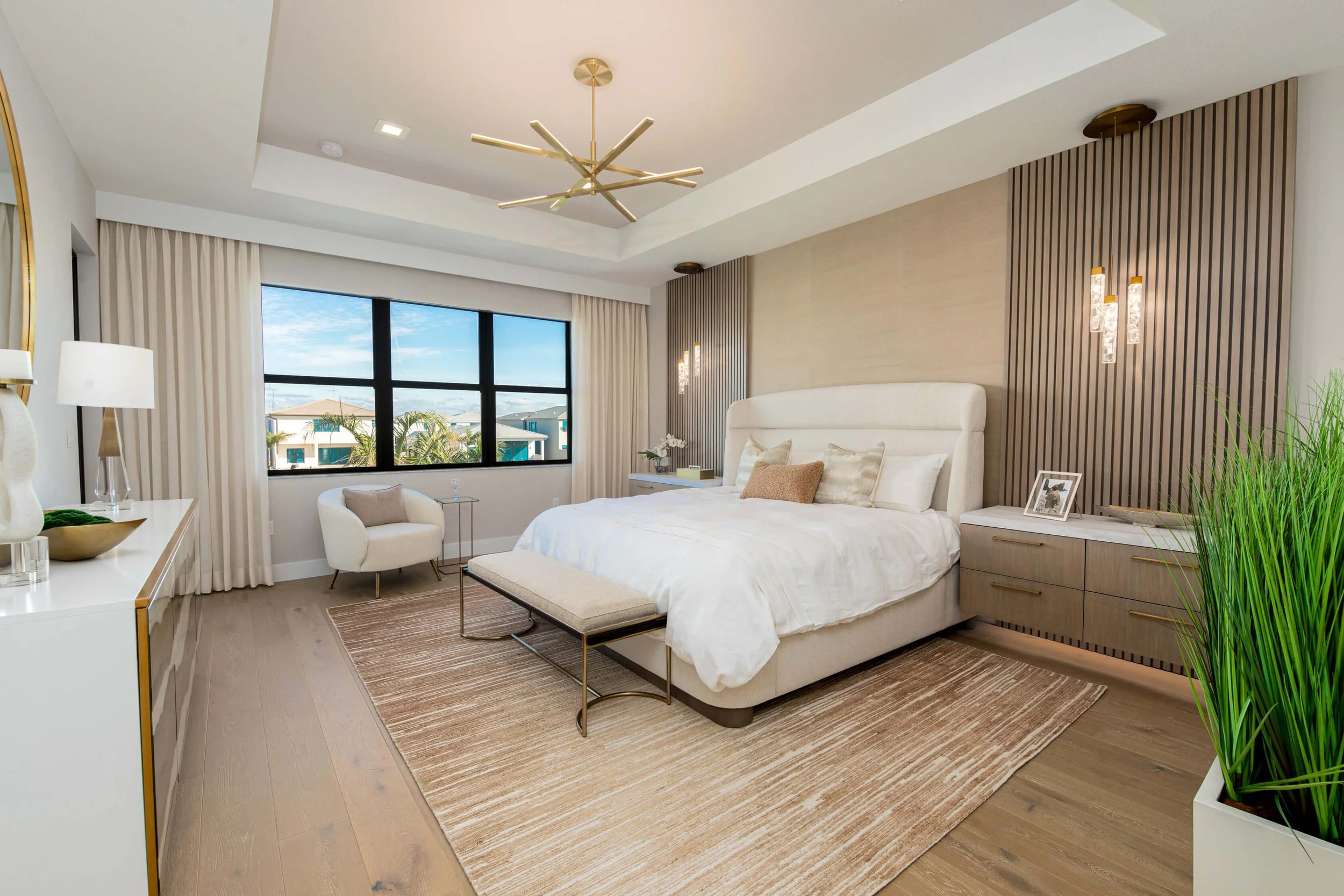 Modern bedroom with king-sized bed, white bedding, beige headboard, and decorative pillows. Light wood floors, a beige rug, a white chair by the window, and built-in nightstands with contemporary light fixtures. Large window with city and ocean view,