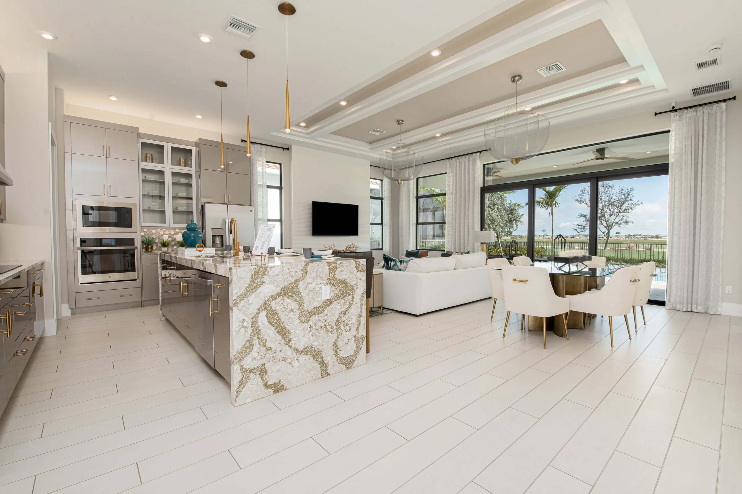 Open-concept kitchen and living area with large windows and sliding glass doors showing an outdoor view of trees and a pool, modern light fixtures, white and gray cabinetry, marble island, white sofa, round dining table with white chairs.