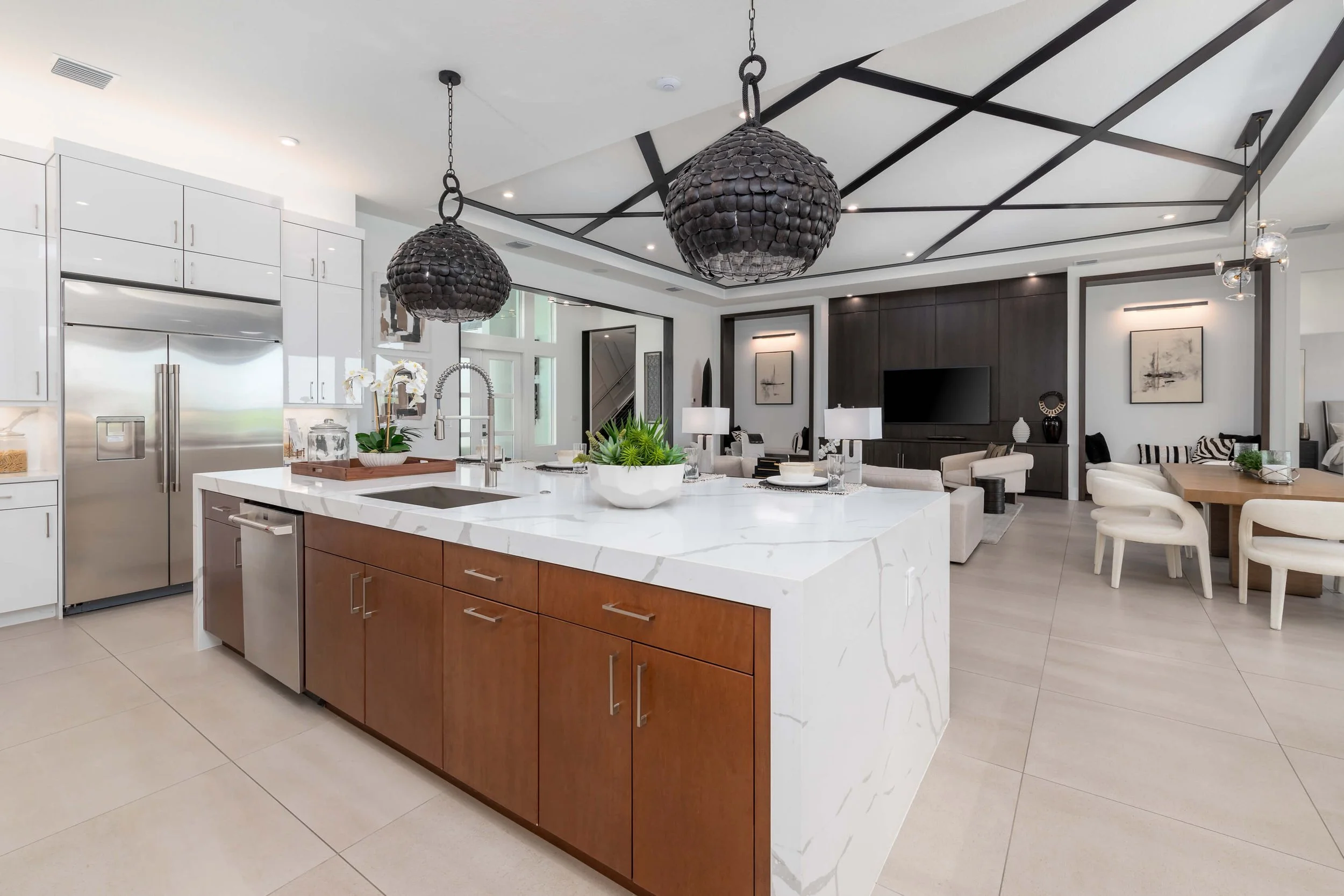 Modern open-concept kitchen and living area with white cabinets, a large marble kitchen island with wooden cabinets, black pendant lights, and a living room with a wall-mounted TV and comfortable seating.