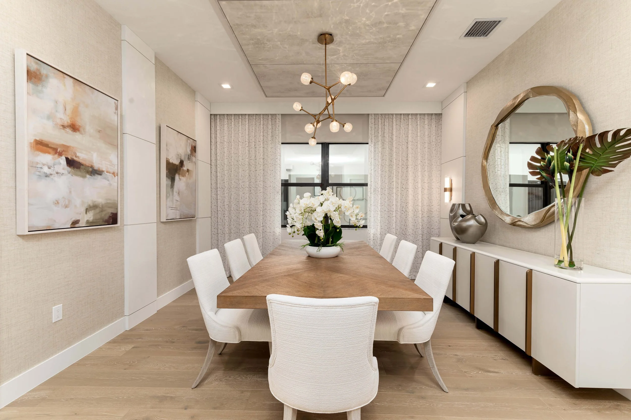 Modern dining room with a wooden table, white upholstered chairs, abstract artwork on beige walls, a large mirror, decorative vases, and a flower arrangement on the table. Beige curtains and a contemporary chandelier are also visible.