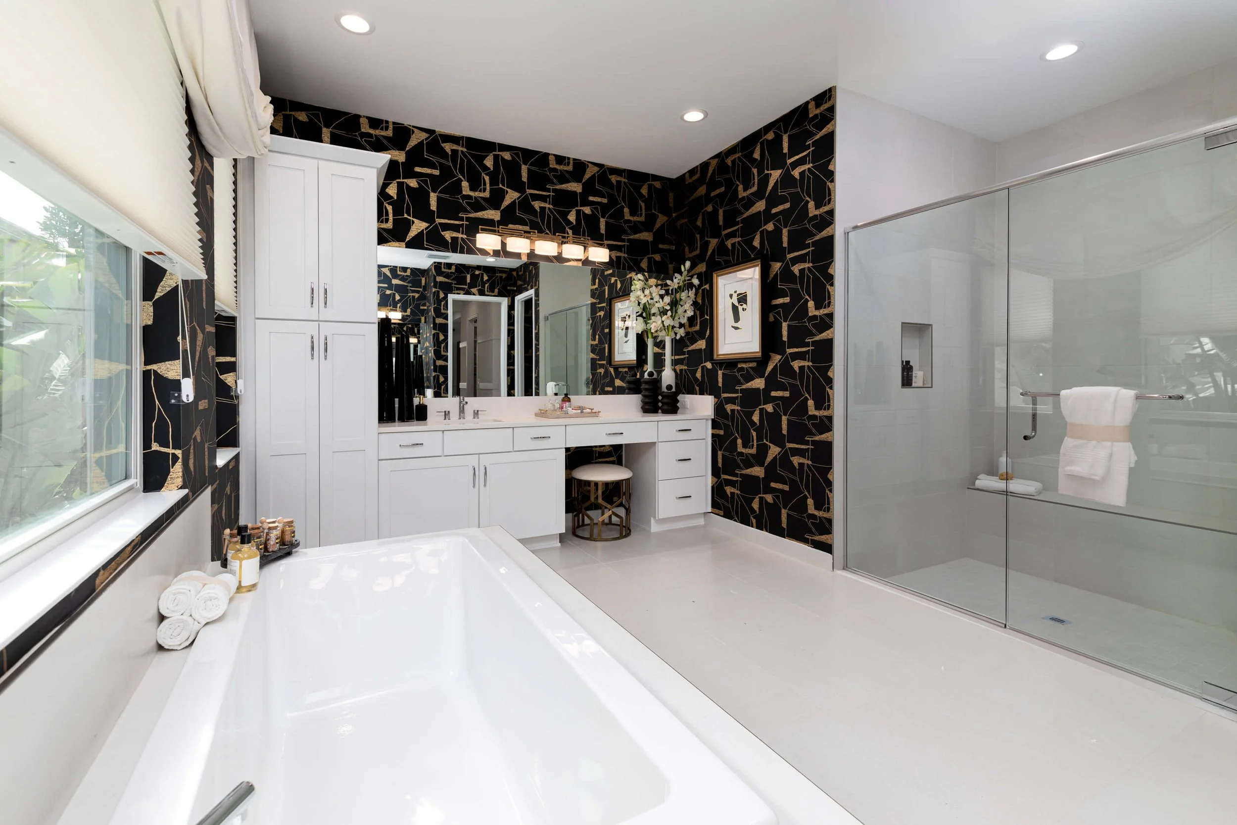 Modern bathroom with black and gold patterned accent wall, white cabinetry, bathtub, glass shower enclosure, and decorative artwork.