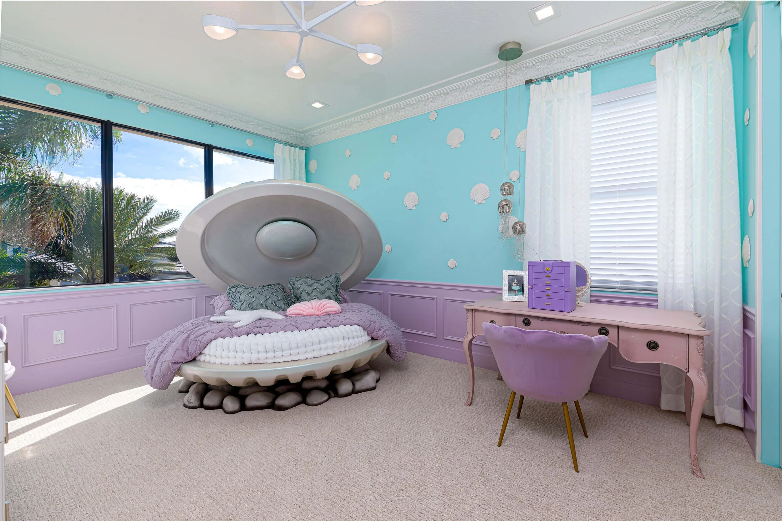 A children's bedroom with a shell-themed decor, including a bed with a shell-shaped headboard, purple and white bedding, and sea shell wall decorations. There is a pink vintage style desk with a purple chair, a framed photo, and a purple letter organ