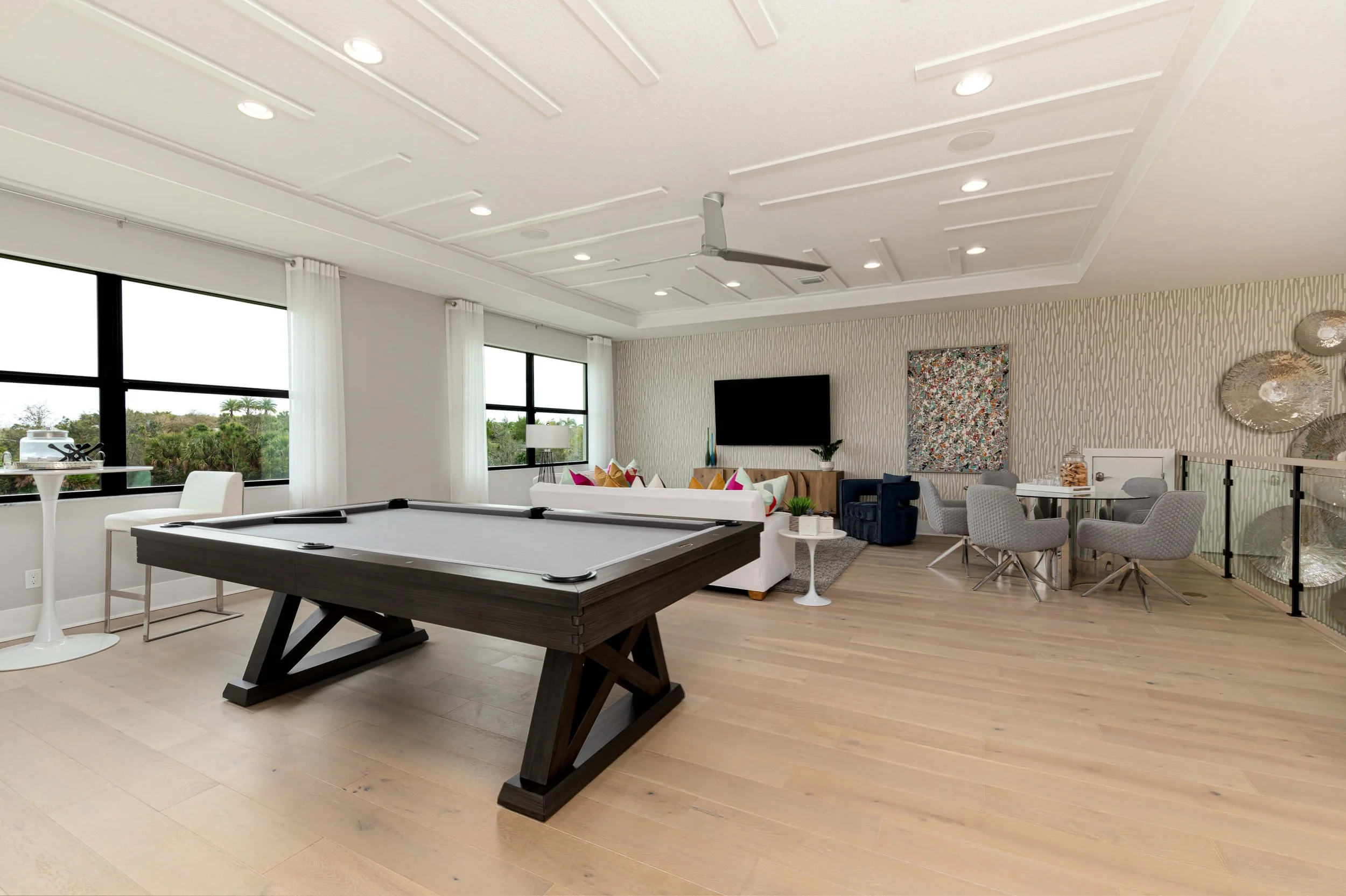 Modern living room with a pool table, large windows, a white sofa with colorful pillows, a wall-mounted TV, a round dining table with four chairs, and decorative wall art.