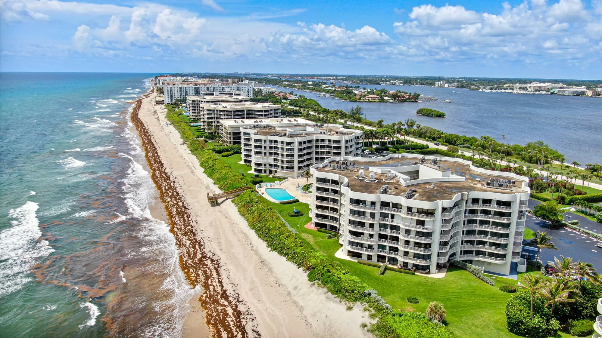 Aerial view of beachfront high-rise condominiums along a sandy shoreline with ocean waves, lush greenery, and a waterway in the background under a partly cloudy sky.