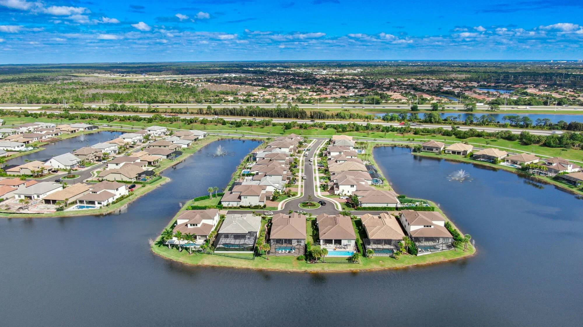 Aerial view of a suburban residential community with houses, water features, and fountains, surrounded by lush green landscapes and a distant cityscape under a blue sky.
