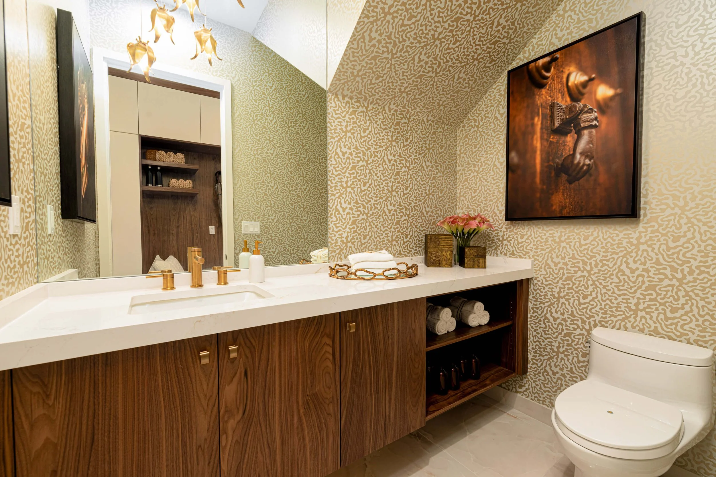 Modern bathroom with a large mirror, wooden vanity with gold fixtures, rolled towels, pink flowers, and a wall-mounted TV. Patterned wallpaper and a white toilet are visible.