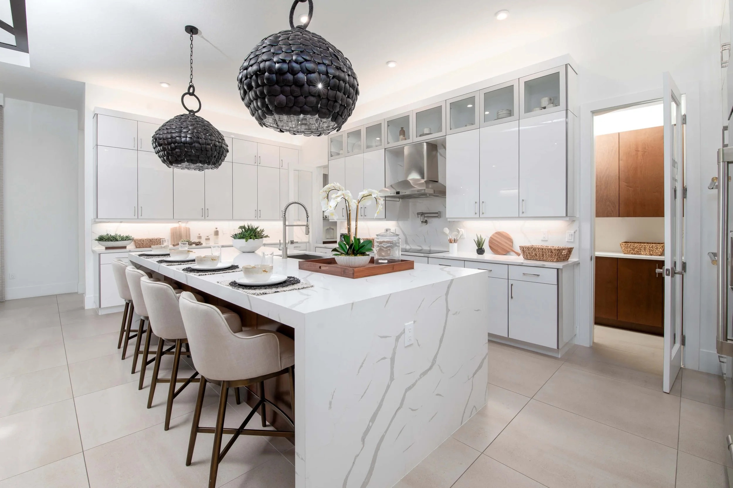 Modern white kitchen with marble island, beige chairs, black pendant lights, and minimal decor.