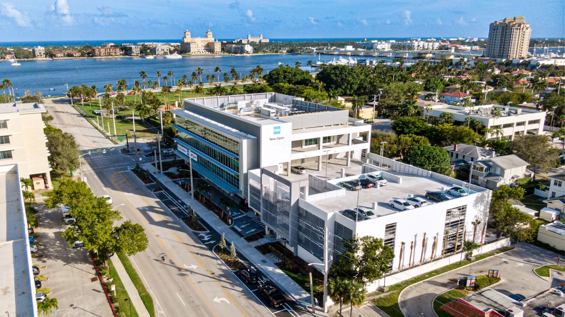 Aerial view of a modern white building with parking lots, surrounded by residential houses, trees, a waterway, and distant buildings in a sunny cityscape.