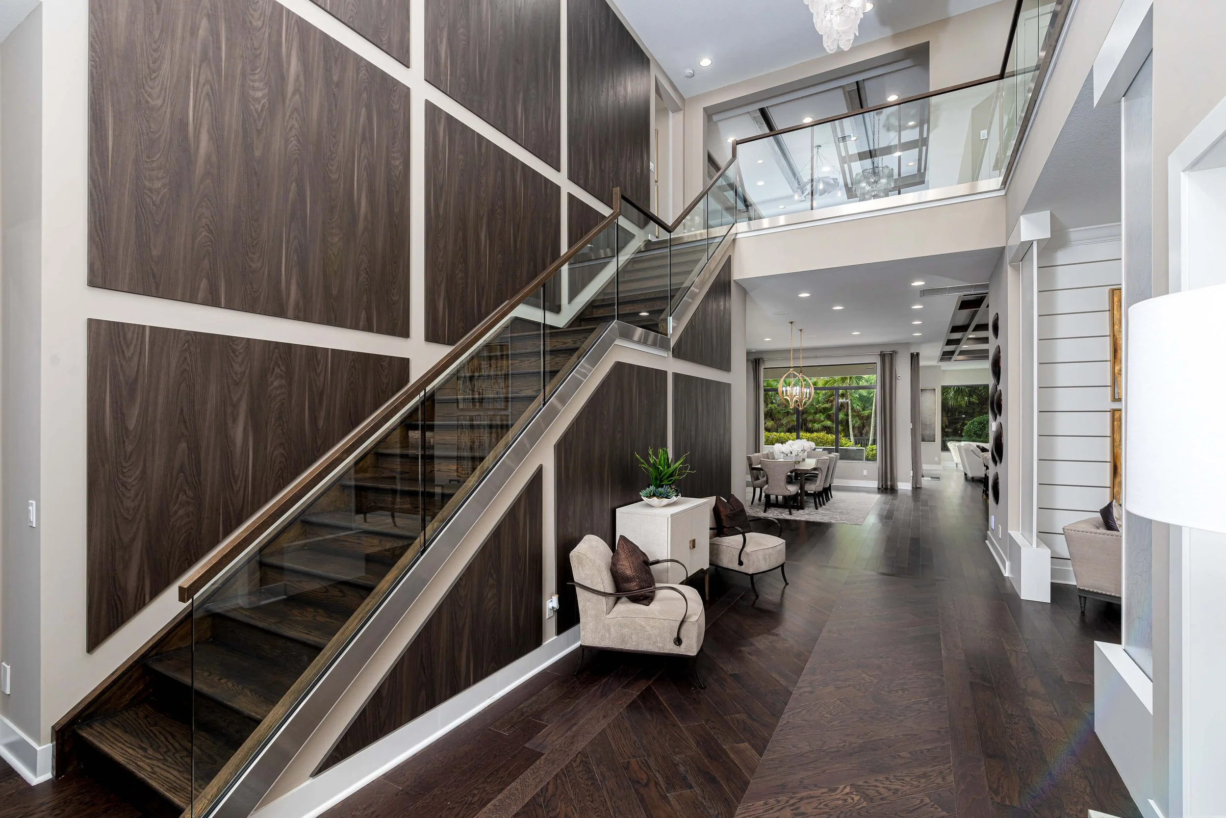 Interior of a modern home with a staircase, dark wood flooring, and a view into a dining area with large windows and view of greenery outside.