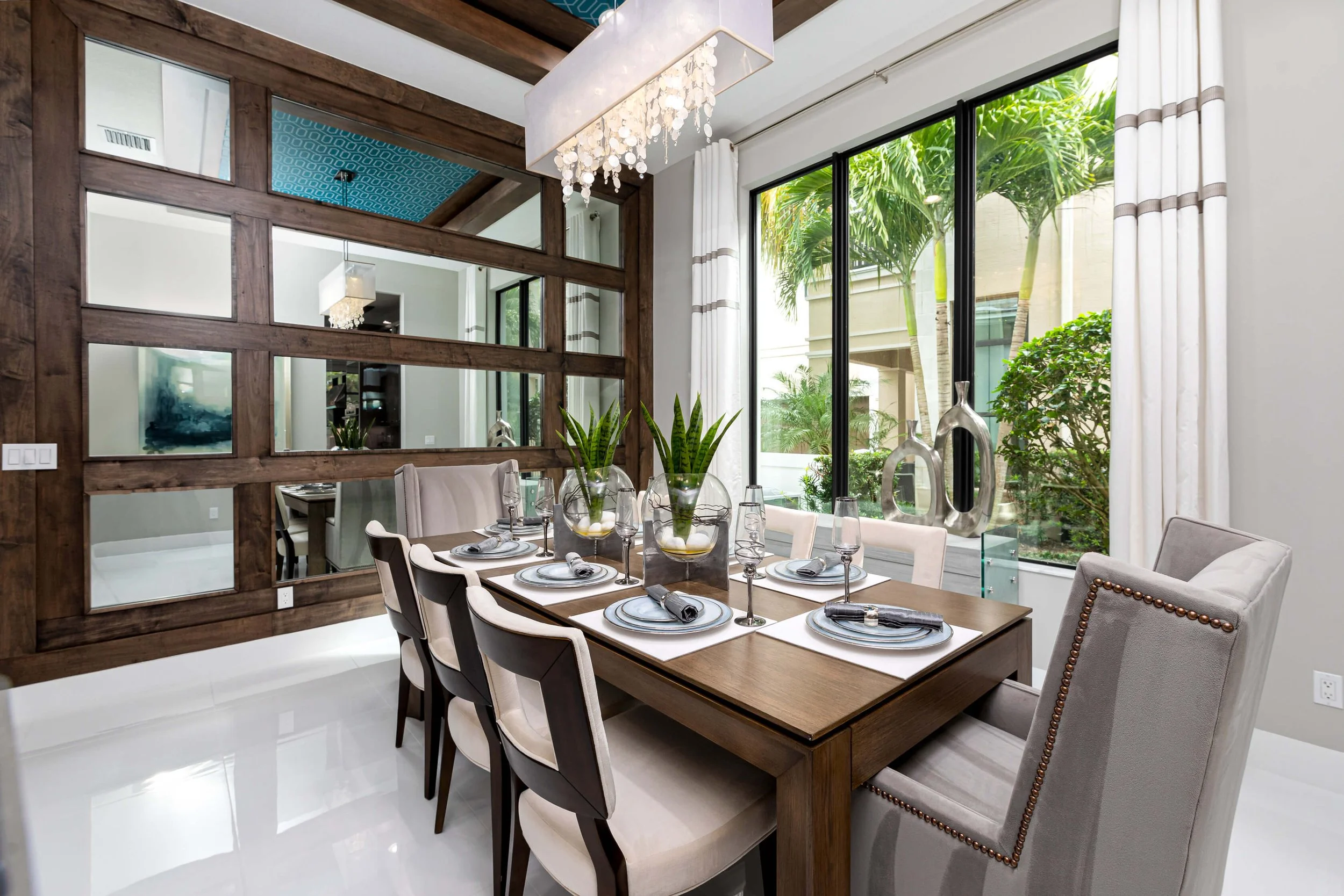 Dining room with a wooden table set with plates, glasses, napkins, and potted plants, next to large windows with white curtains showing outdoor trees and bushes.