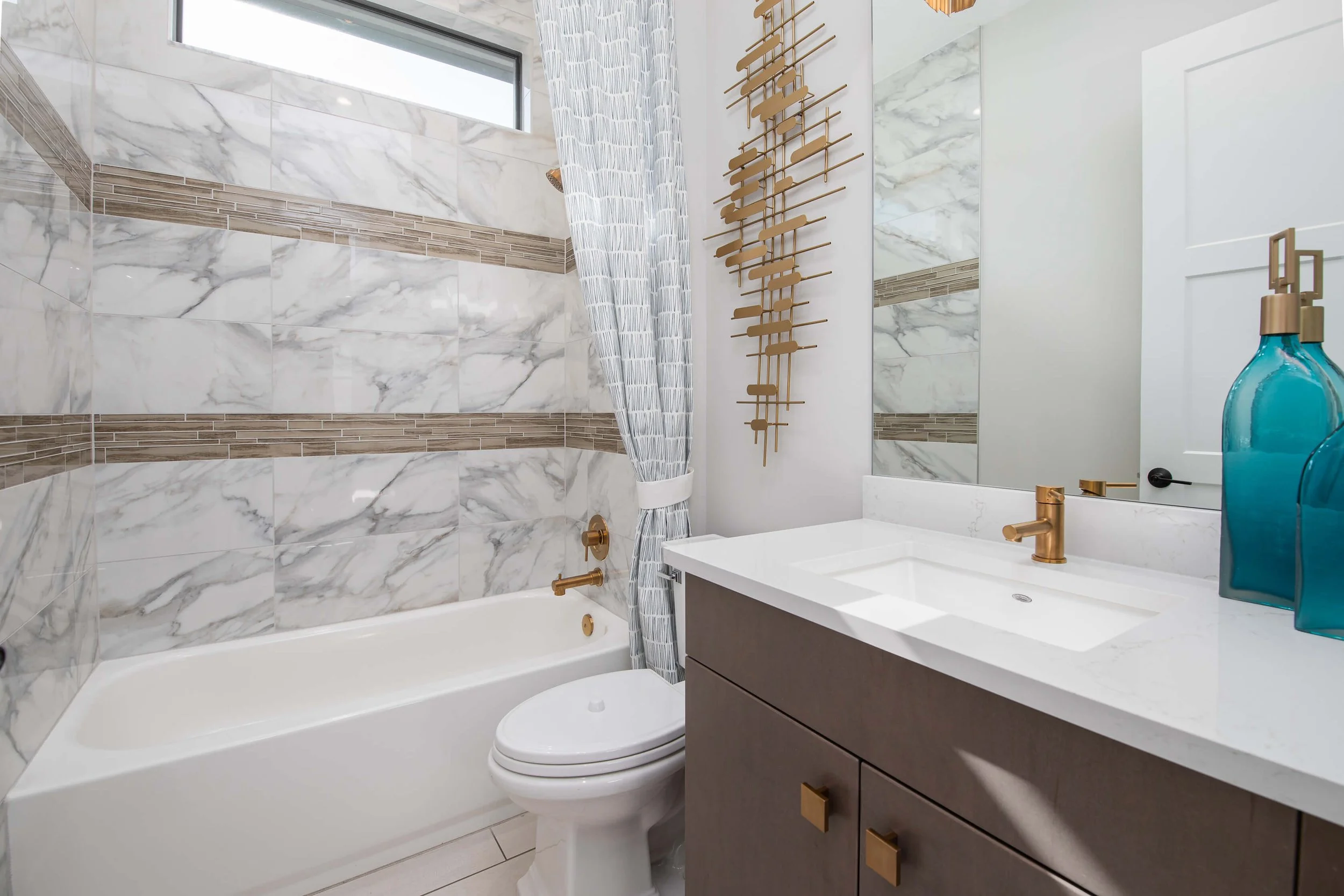 Modern bathroom with marble tiled walls, a bathtub with gold fixtures, a toilet, a vanity with a white countertop, gold faucet, and a mirror. Decor includes blue glass bottles and a gold wall art piece.