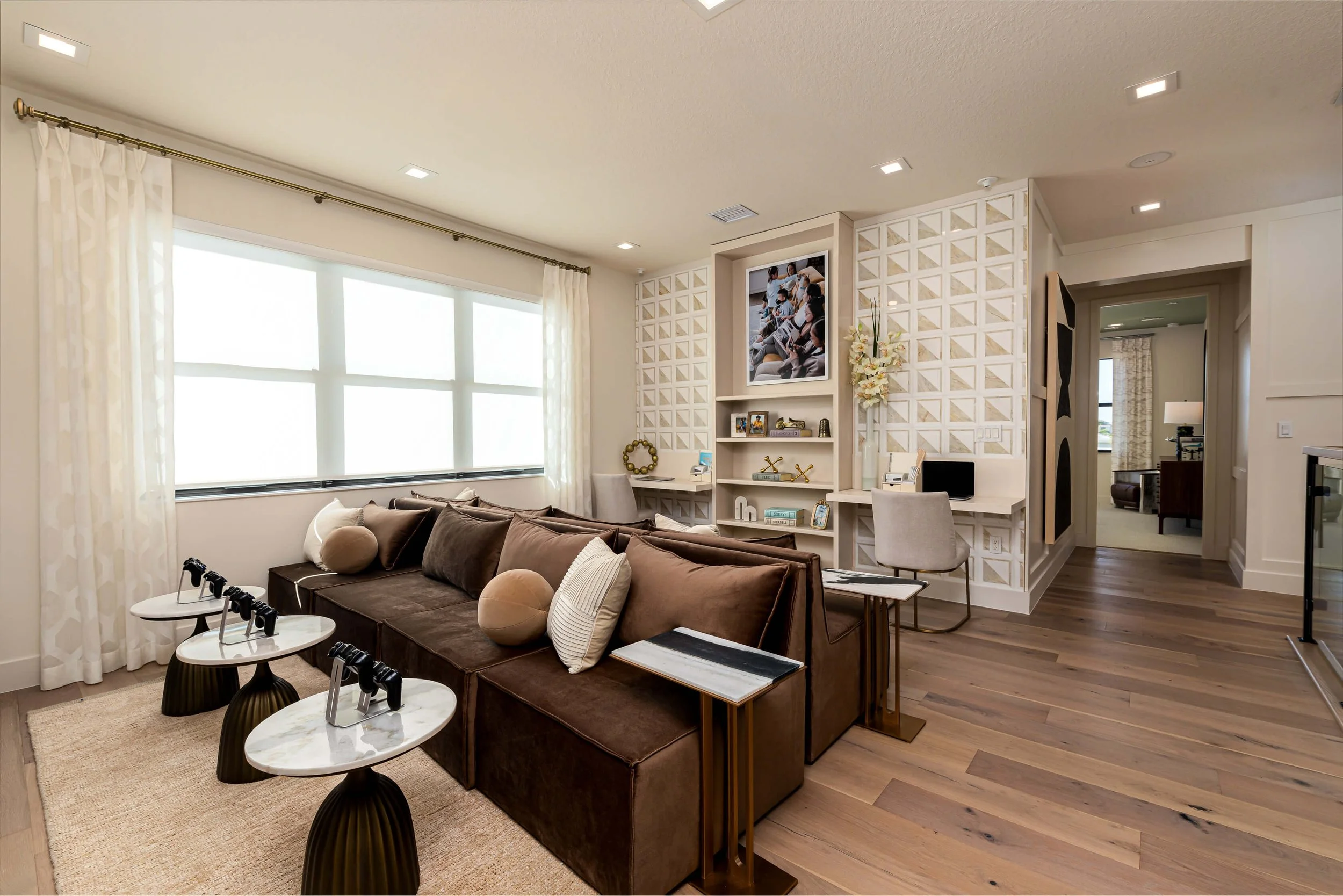 Modern living room with large window, brown sofa with pillows, white curtains, decorative shelves, TV, and a desk with chair.