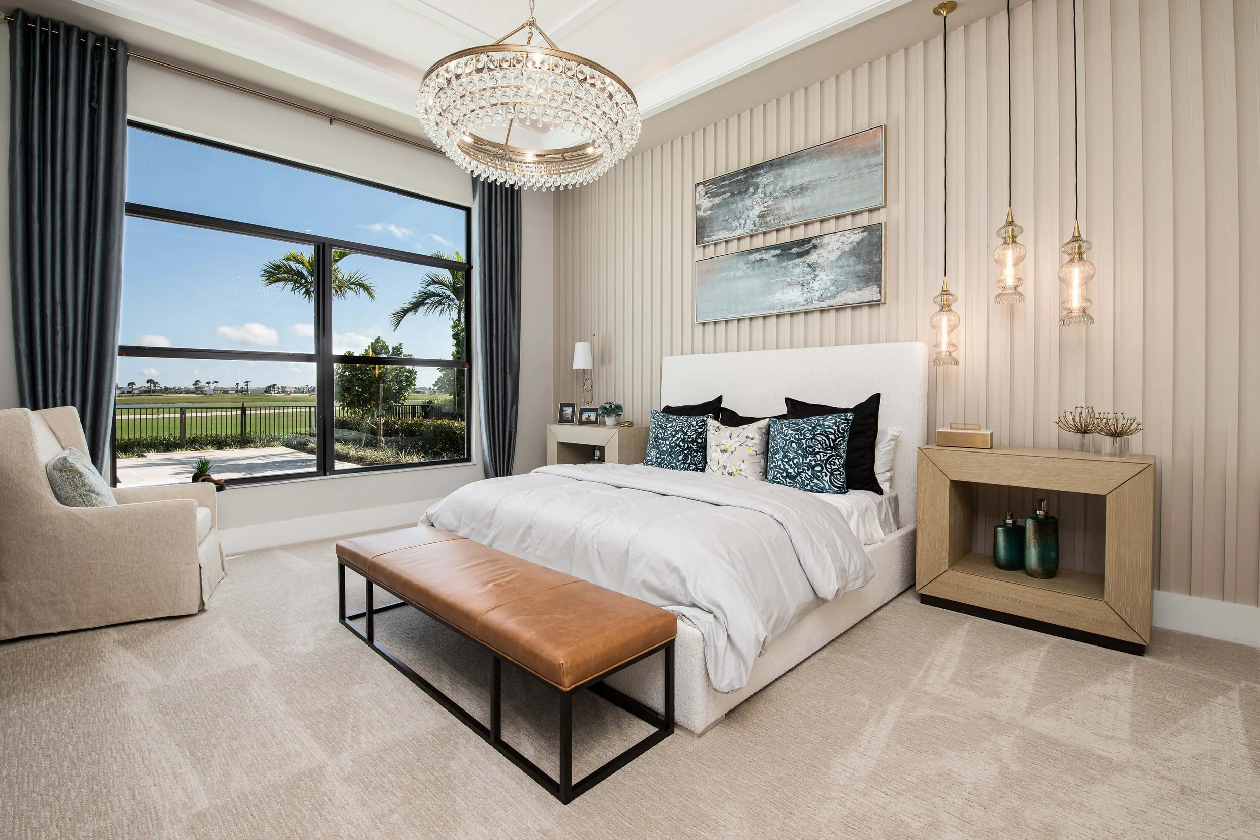 Modern bedroom with large window, white bed with multiple pillows, beige carpet, chandelier, and contemporary decor.