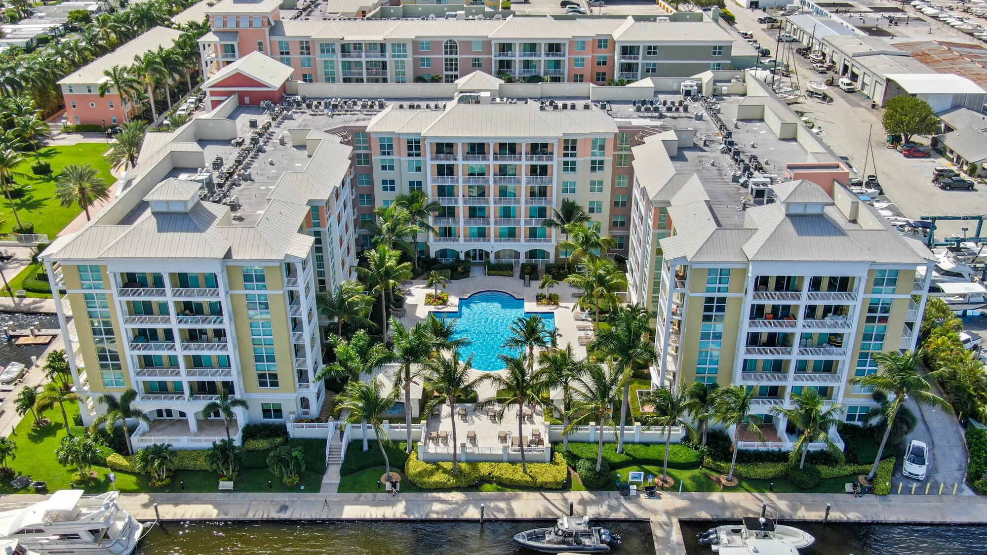 Aerial view of a multi-story residential building complex with a swimming pool, palm trees, and waterway with boats docked.