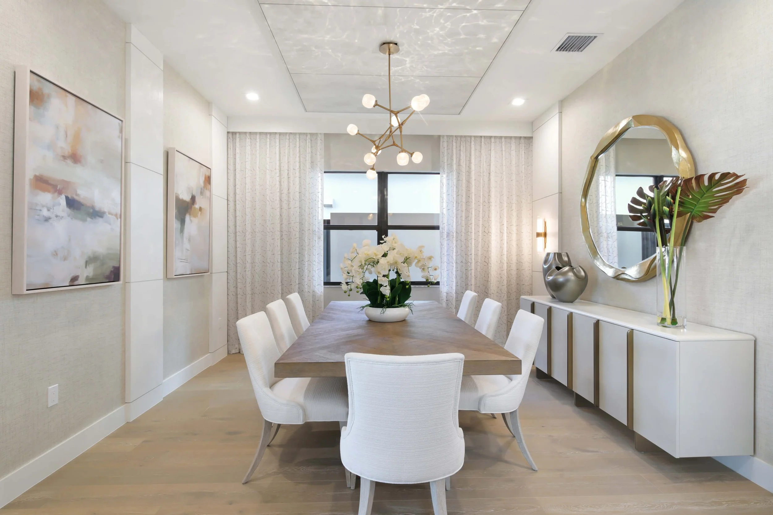 Modern dining room with a wooden table surrounded by eight white upholstered chairs, a large vase with white orchids in the center, abstract paintings on the wall, a gold-framed mirror, decorative vases, and a large window with white curtains.
