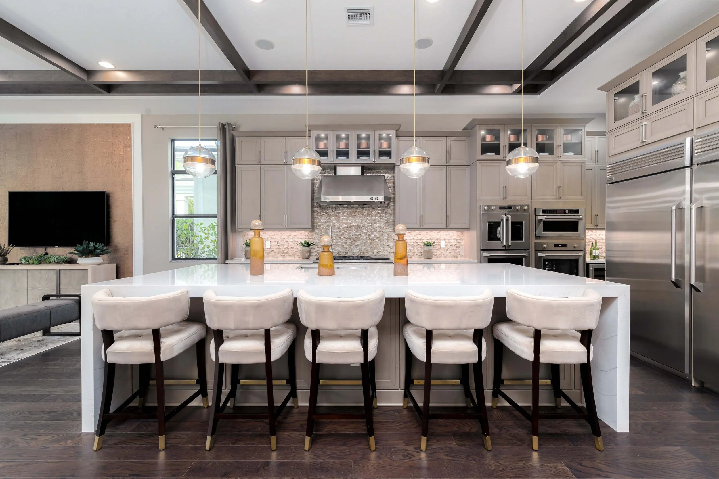 Modern kitchen with light gray cabinets, a large white island with seven upholstered barstools, pendant lights, stainless steel appliances, and a dark wood floor.