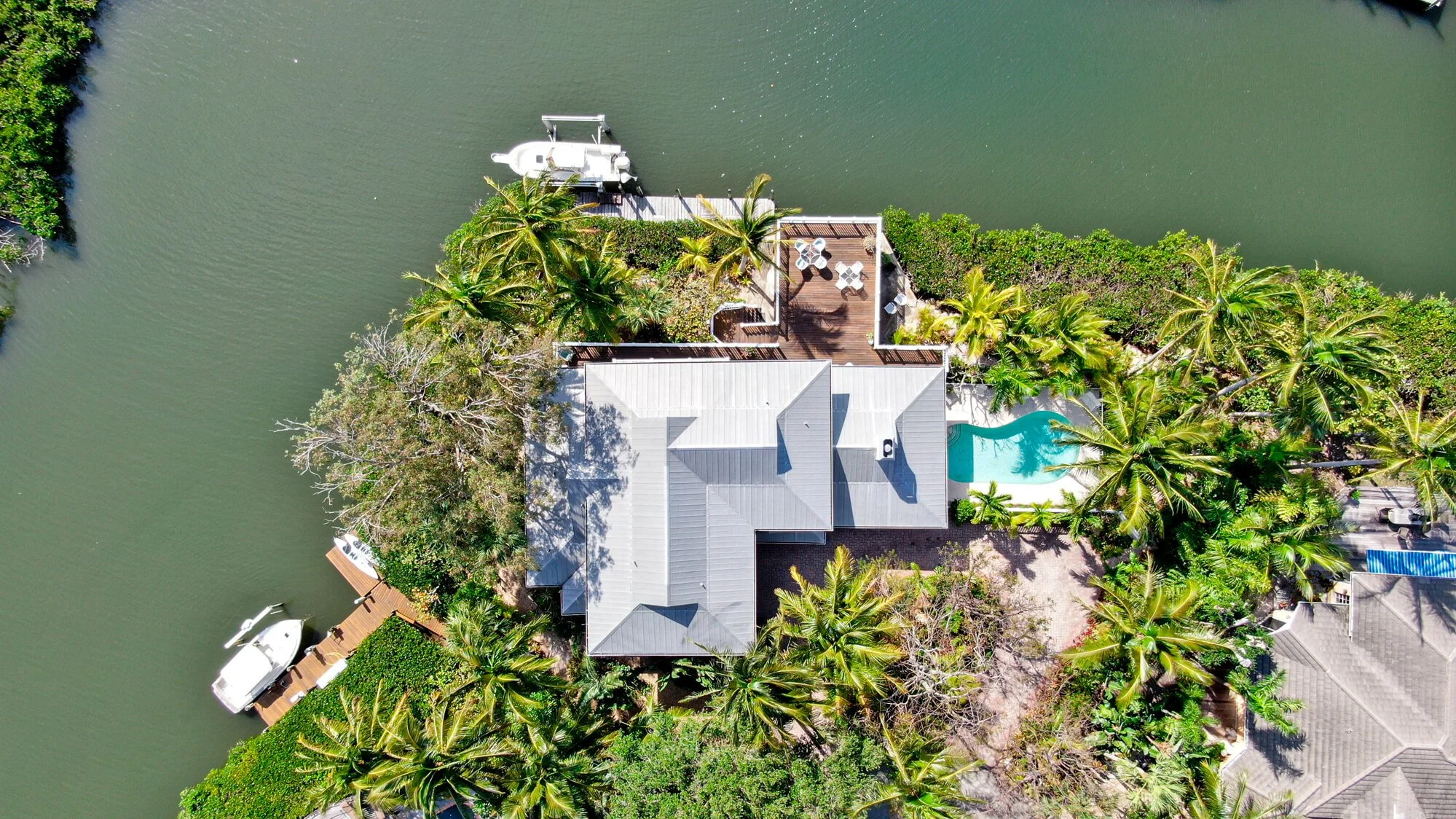 Aerial view of a house with a metal roof, surrounded by palm trees, near a waterway with boats and a dock, and a swimming pool in the backyard.