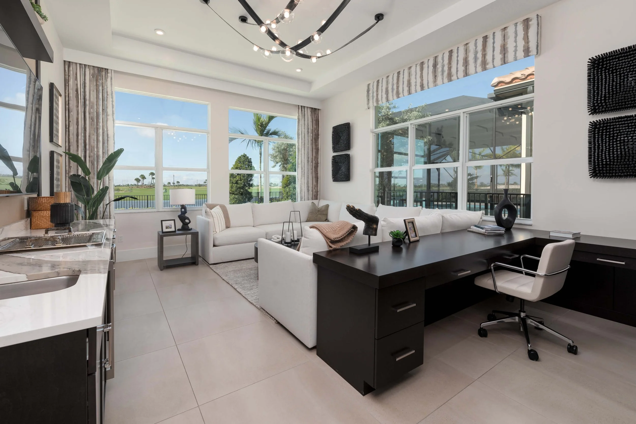 Modern living room with large windows, white sectional sofa, black desk with white chair, and outdoor view of trees and blue sky
