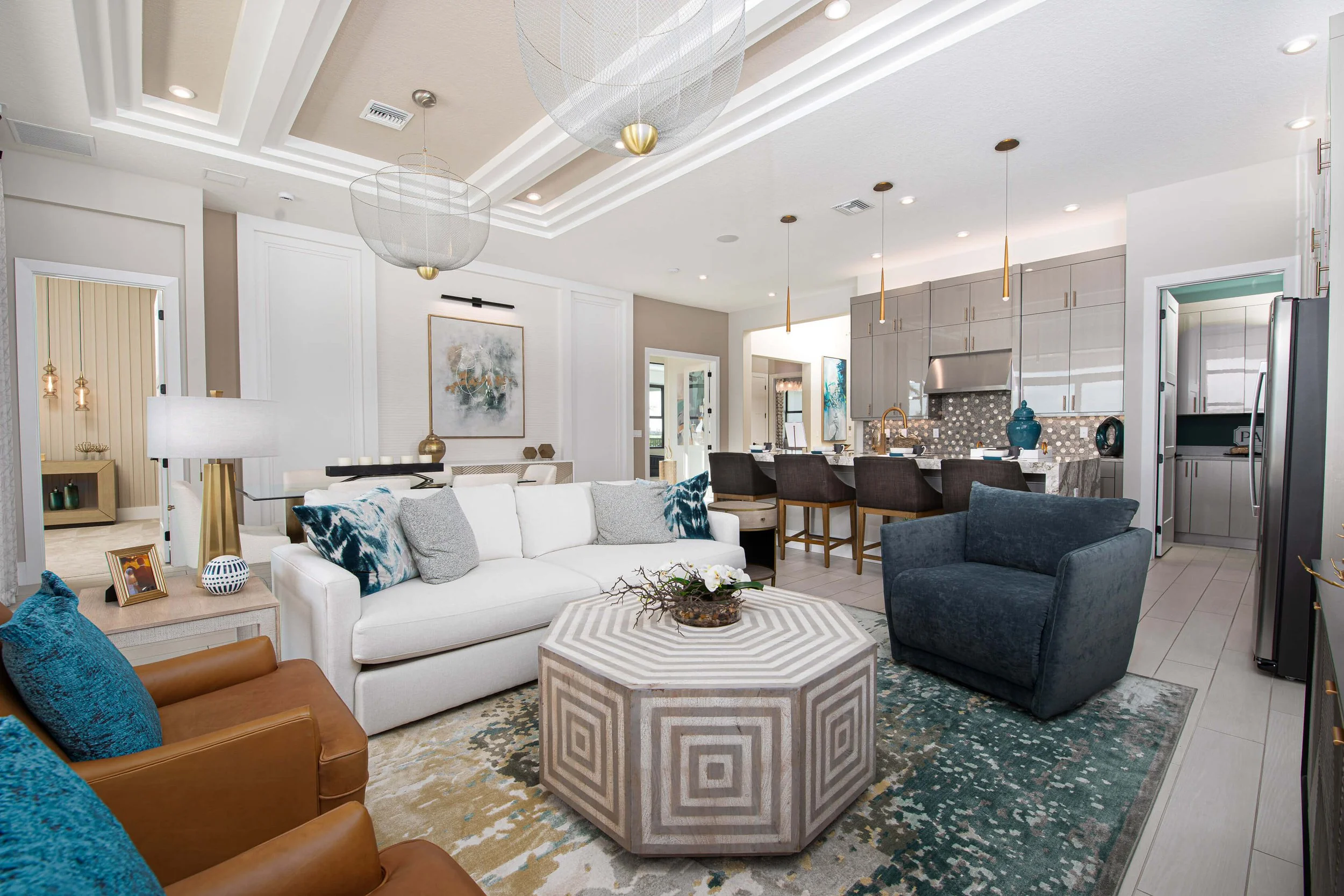 Modern open-concept living room and kitchen with white sofa, blue and gray accent pillows, a geometric coffee table, bar stools at kitchen island, stainless steel appliances, and contemporary lighting.