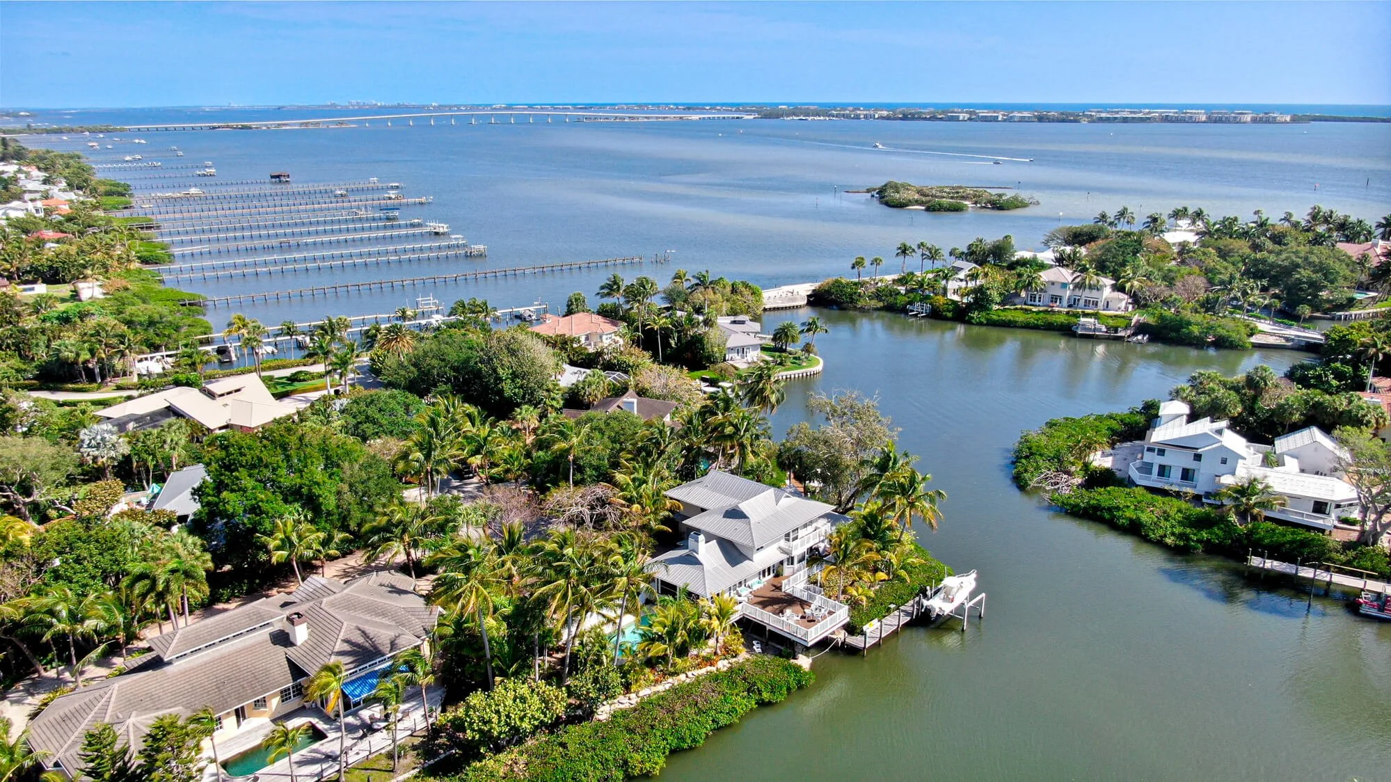 Aerial view of a coastal residential area with houses surrounded by palm trees, a waterway with boats, and a large body of water in the background.