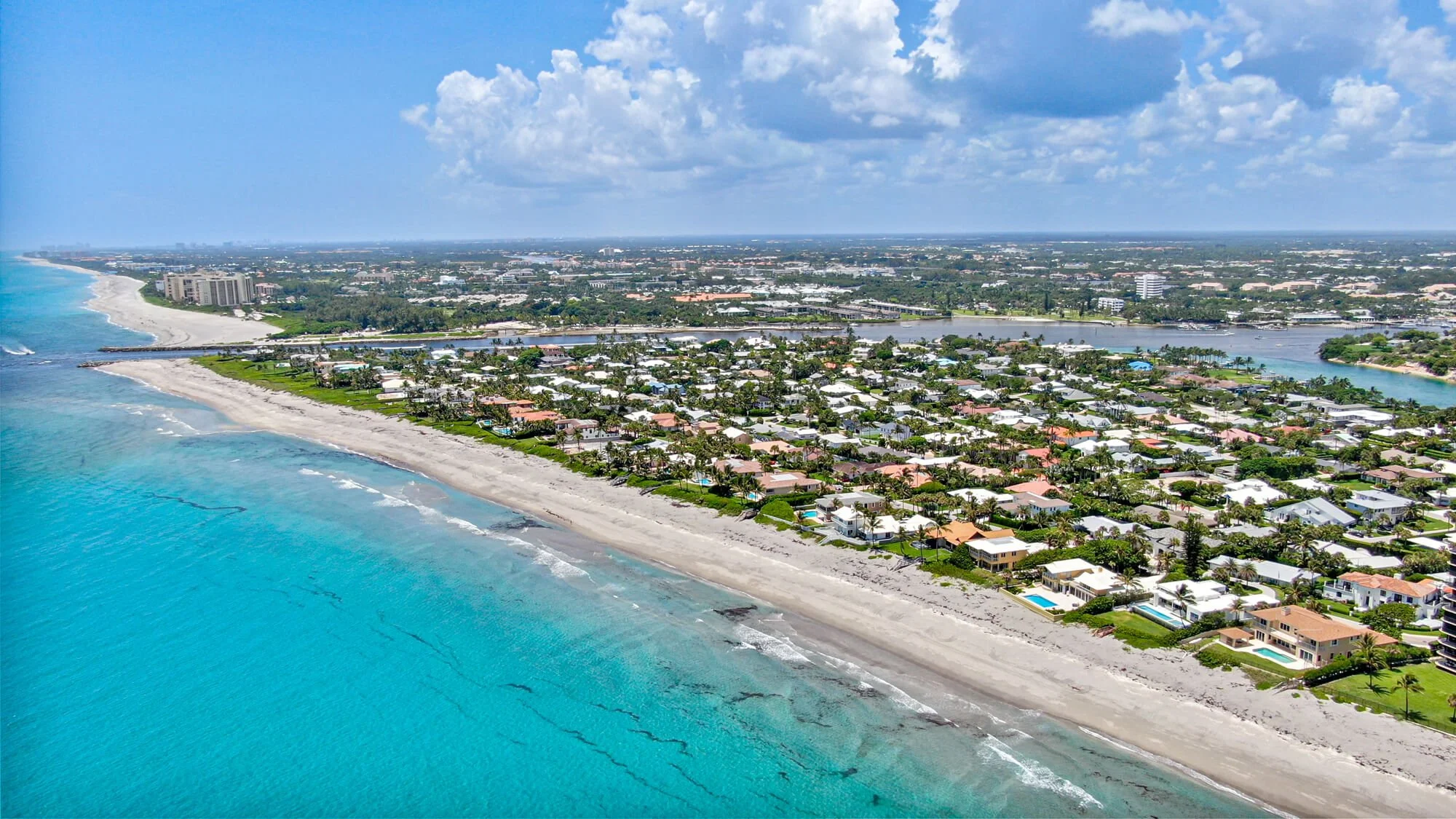 Aerial view of a coastal neighborhood with sandy beach, blue ocean, residential houses, and a cityscape in the background under a partly cloudy sky.