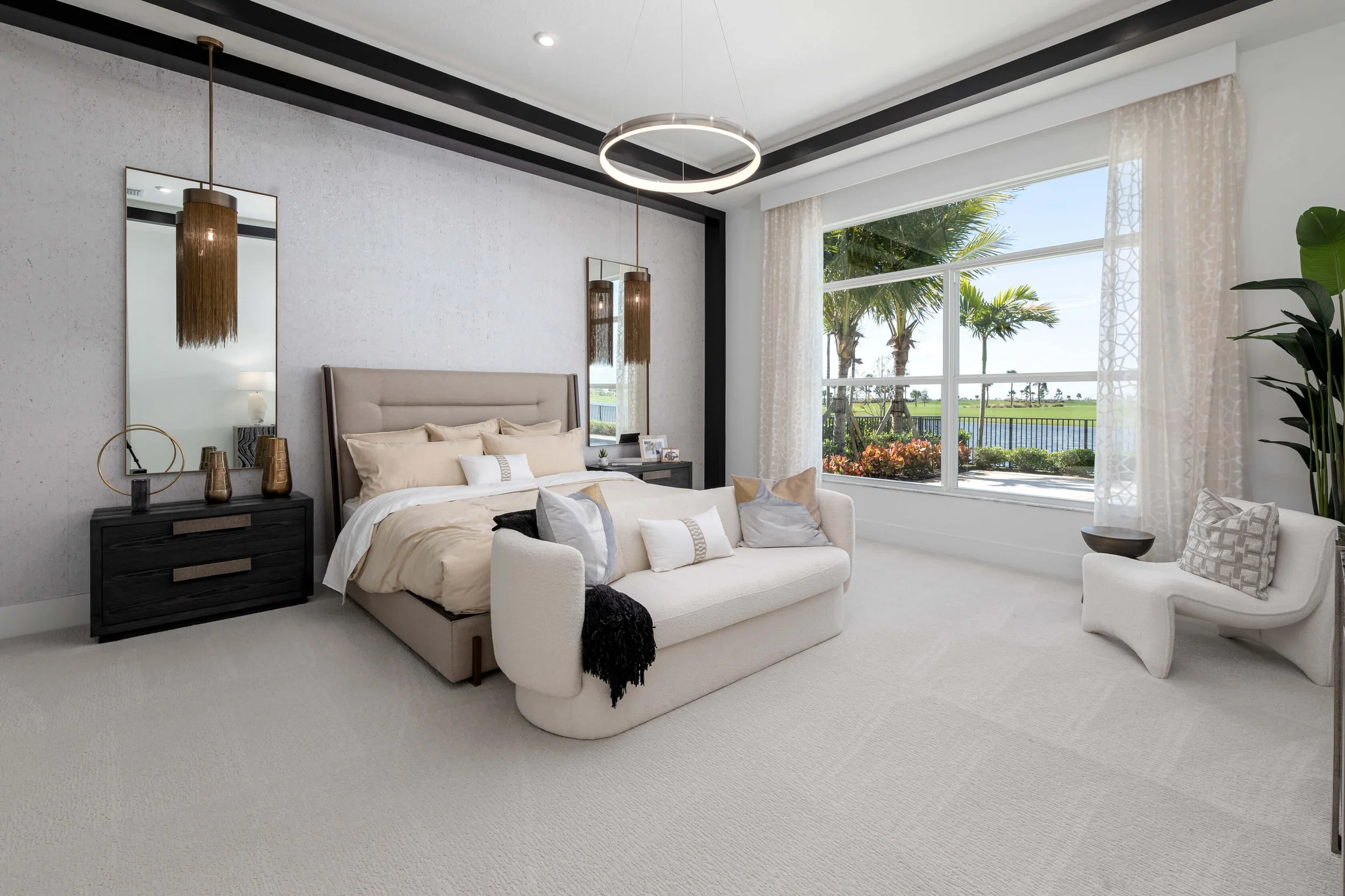 Bright bedroom with large window showing palm trees and water outside, beige bed with pillows, white sofa with pillows, white accent chair, black nightstands, round ceiling light, hanging pendant lights, decorative vases, mirror, and potted plant.